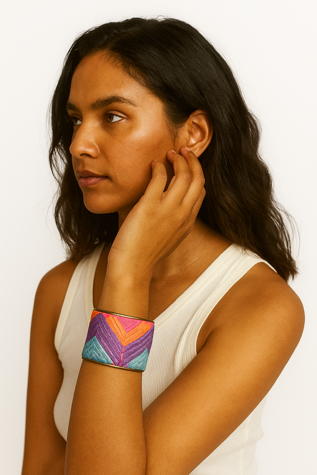 A woman wearing a 2-inch wide brass cuff bracelet wrapped in vibrant woven fabric featuring coral, teal, purple, and pink chevron stripes. Her arm is lifted near her ear, showing the cuff’s shimmer and fit.