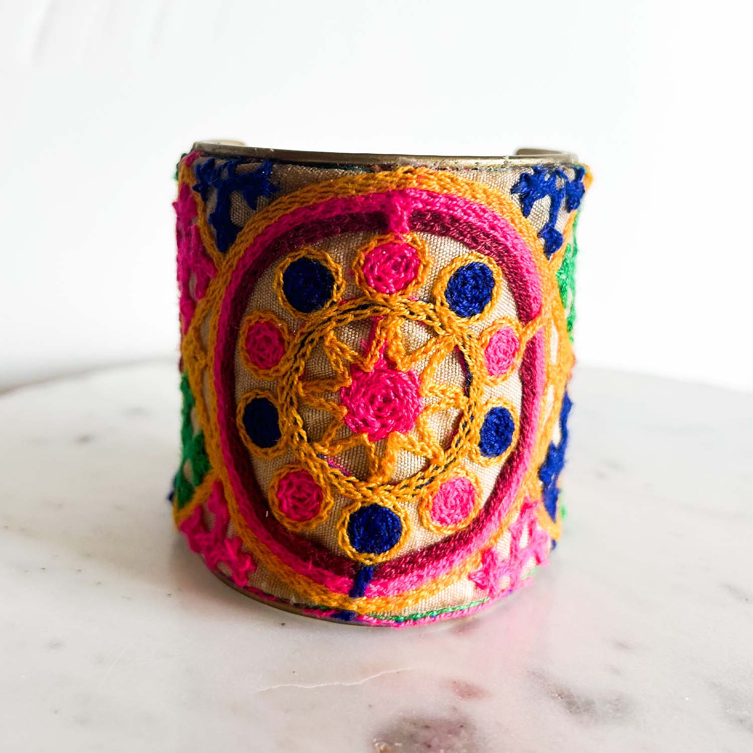 A standalone 3-inch wide raw brass cuff bracelet wrapped in neutral linen fabric, featuring hand-embroidered circular medallion designs in fuchsia, golden orange, emerald green, and cobalt blue threads, photographed front-on against a white background.