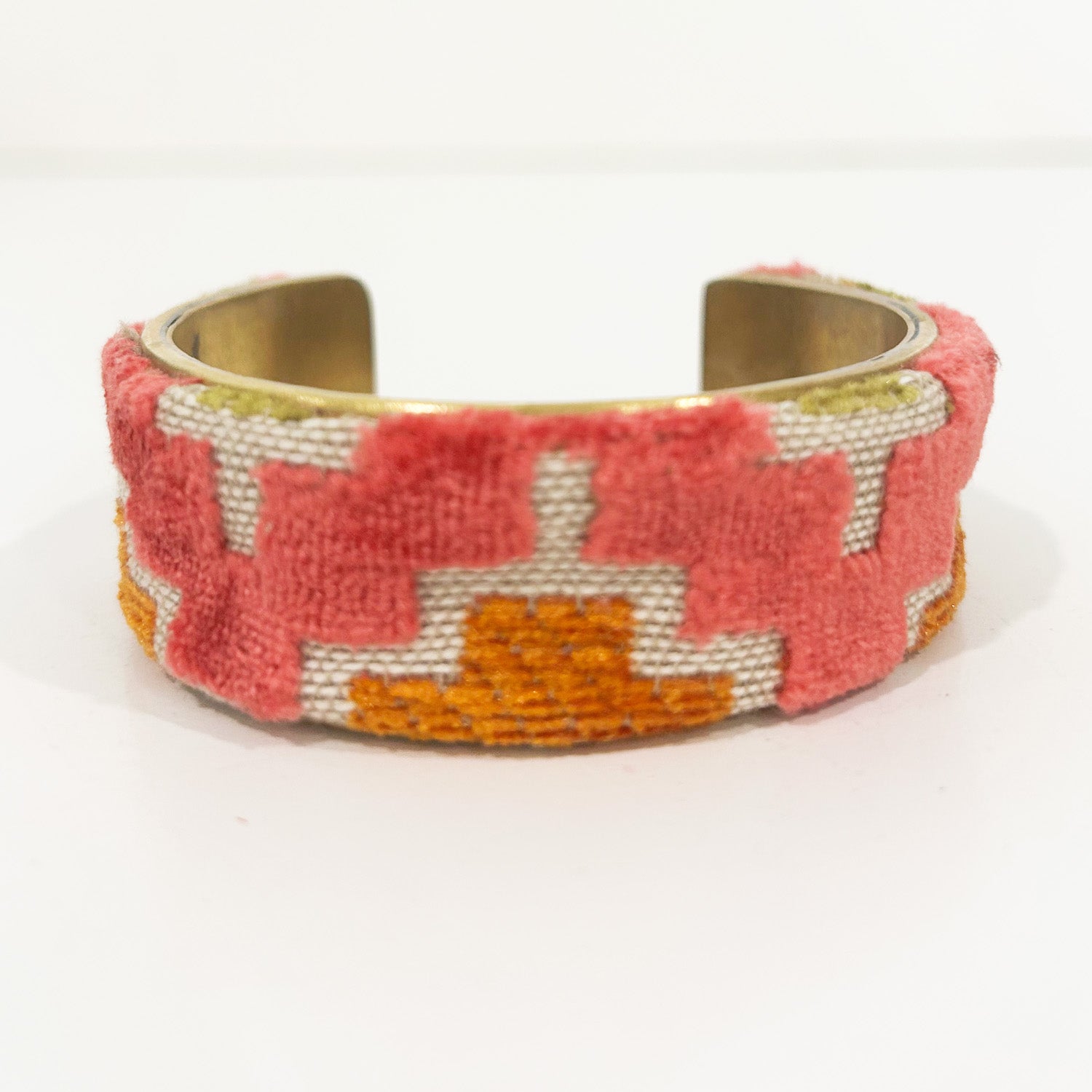 An open-back view of the Theta cuff showing its raw brass interior and velvet exterior with coral and mustard zigzag patterns on neutral linen, placed on a white surface.