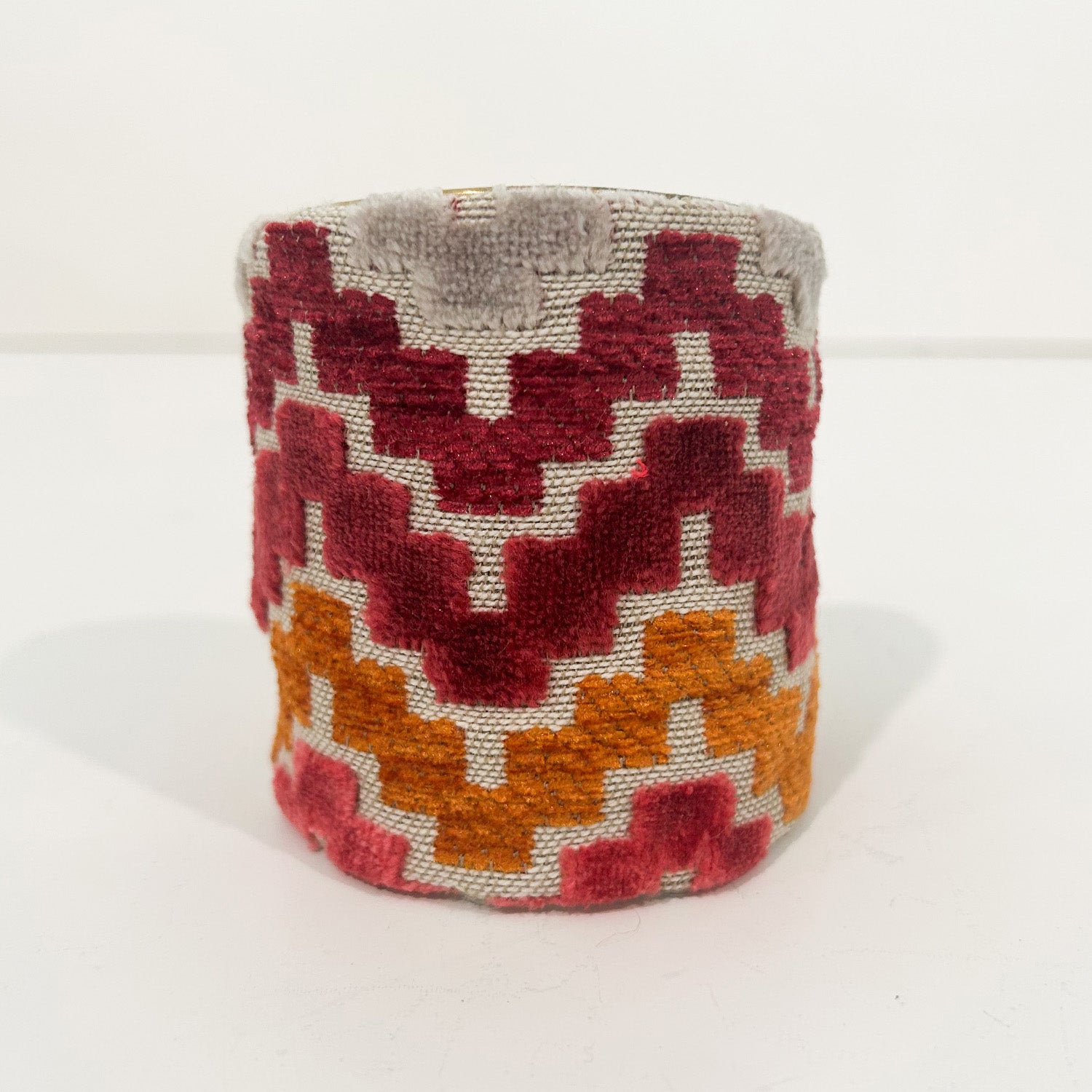 A standalone 3-inch wide raw brass cuff bracelet wrapped in velvet featuring coral, raspberry, and mustard stepped zigzag embroidery on neutral linen, photographed front-on against a white background.