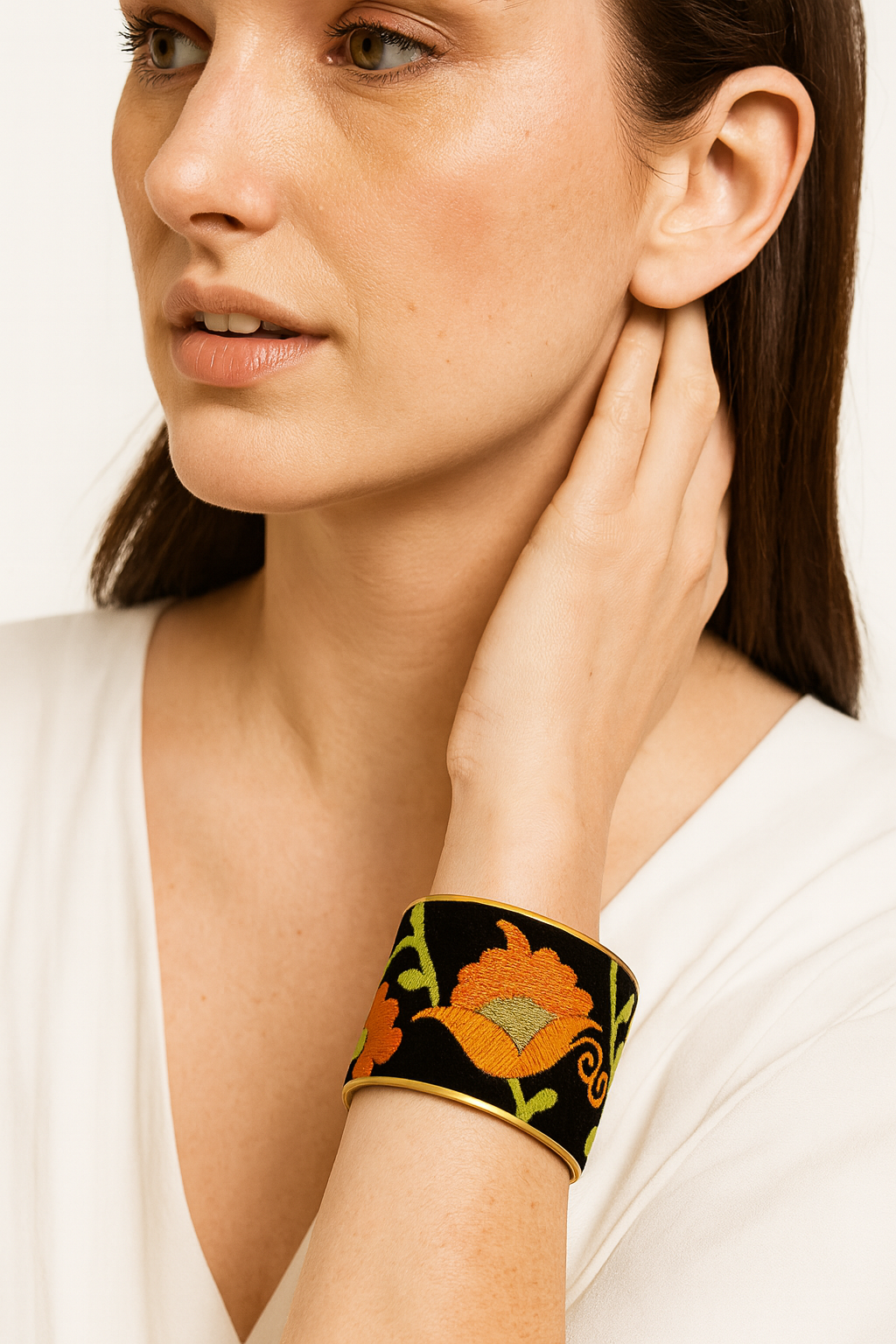 Close-up of a woman’s wrist and lower face, wearing the Redd Dott “Aurora” 3″ wide fabric cuff. The deep black velvet base is embroidered with bold orange poppy blossoms and bright chartreuse-green vines, all set within a gently curved open-back brass frame as her hand lightly touches her hair.