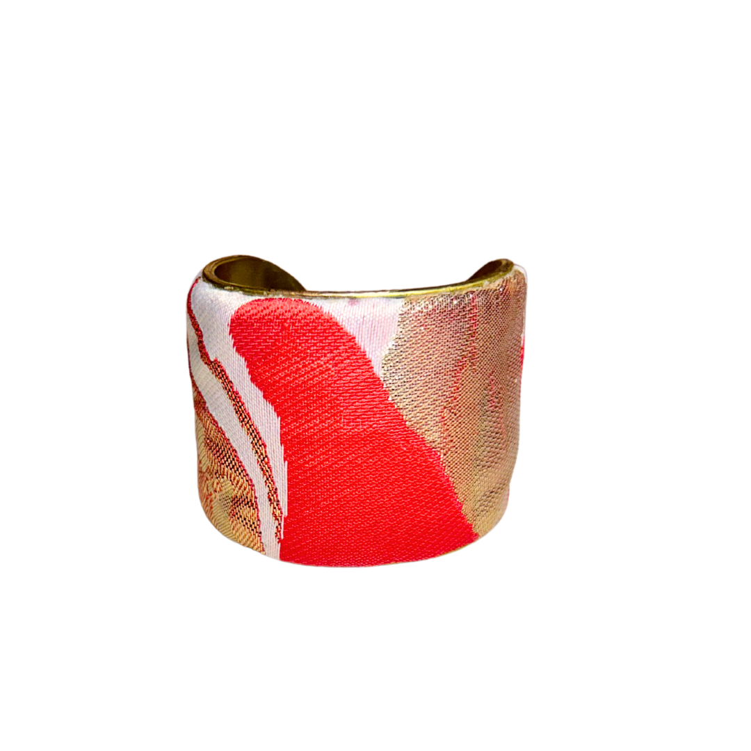A close-up front view of a brass cuff bracelet wrapped in abstract jacquard fabric featuring coral red, metallic gold, and white colorblocks, highlighting the raw brass edges