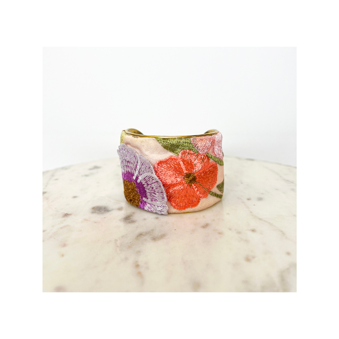 A wide, gold cuff bracelet featuring detailed embroidered flowers in shades of pink, orange, and purple, displayed on a round marble surface. This vintage brass cuff captures the essence of spring with its vibrant floral design.