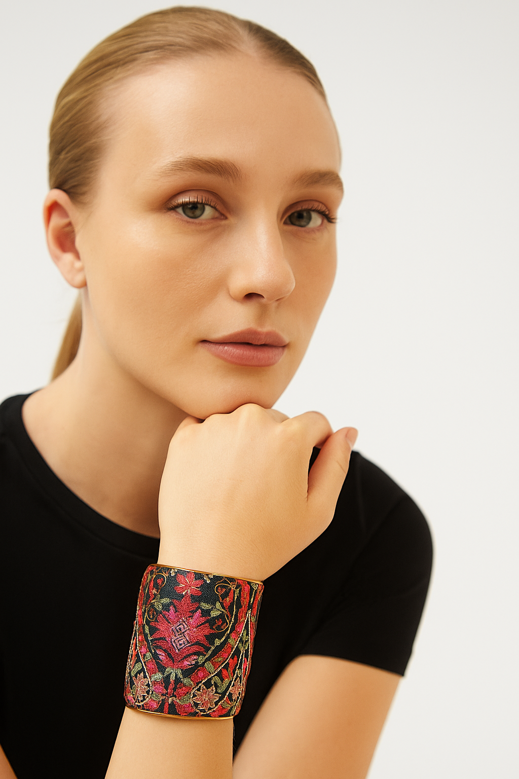 A three-inch brass cuff bracelet wrapped in black hand-embroidered fabric featuring cerise, fuchsia, and olive green floral motifs, shown on a female model wearing a black top; the open-back adjustable fit and the model’s serene expression are both visible.
