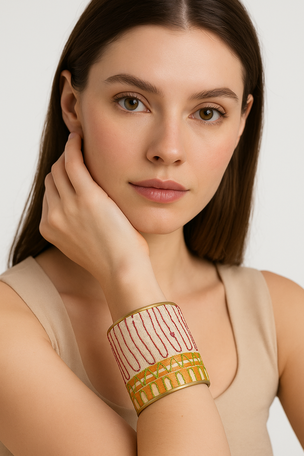 A female model in a beige tank rests her chin on her hand, wearing a three-inch brass cuff bracelet wrapped in cream silk with coral loop stitches and olive-amber embroidery; the open-back adjustable fit is visible against a white background.