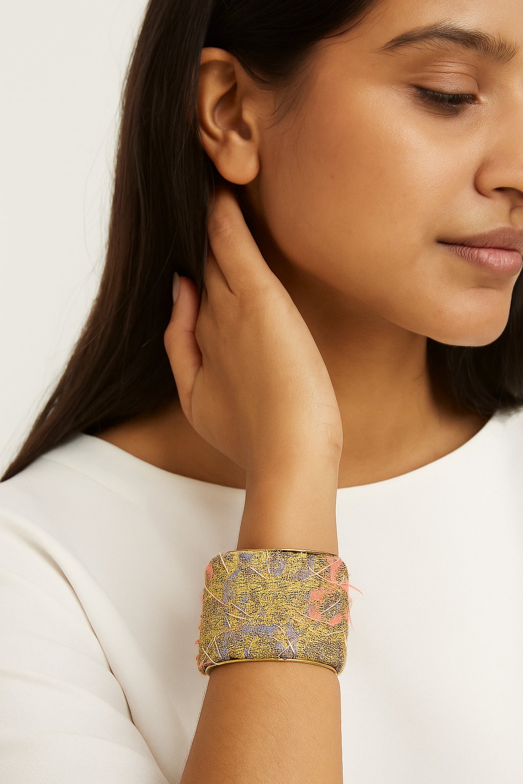 A female model in a white blouse wears a three-inch brass cuff bracelet wrapped in golden jacquard fabric with sparkling crystal beads and a blue accent gem; the adjustable open-back fit and her hand tucked near her ear are visible against a neutral background.