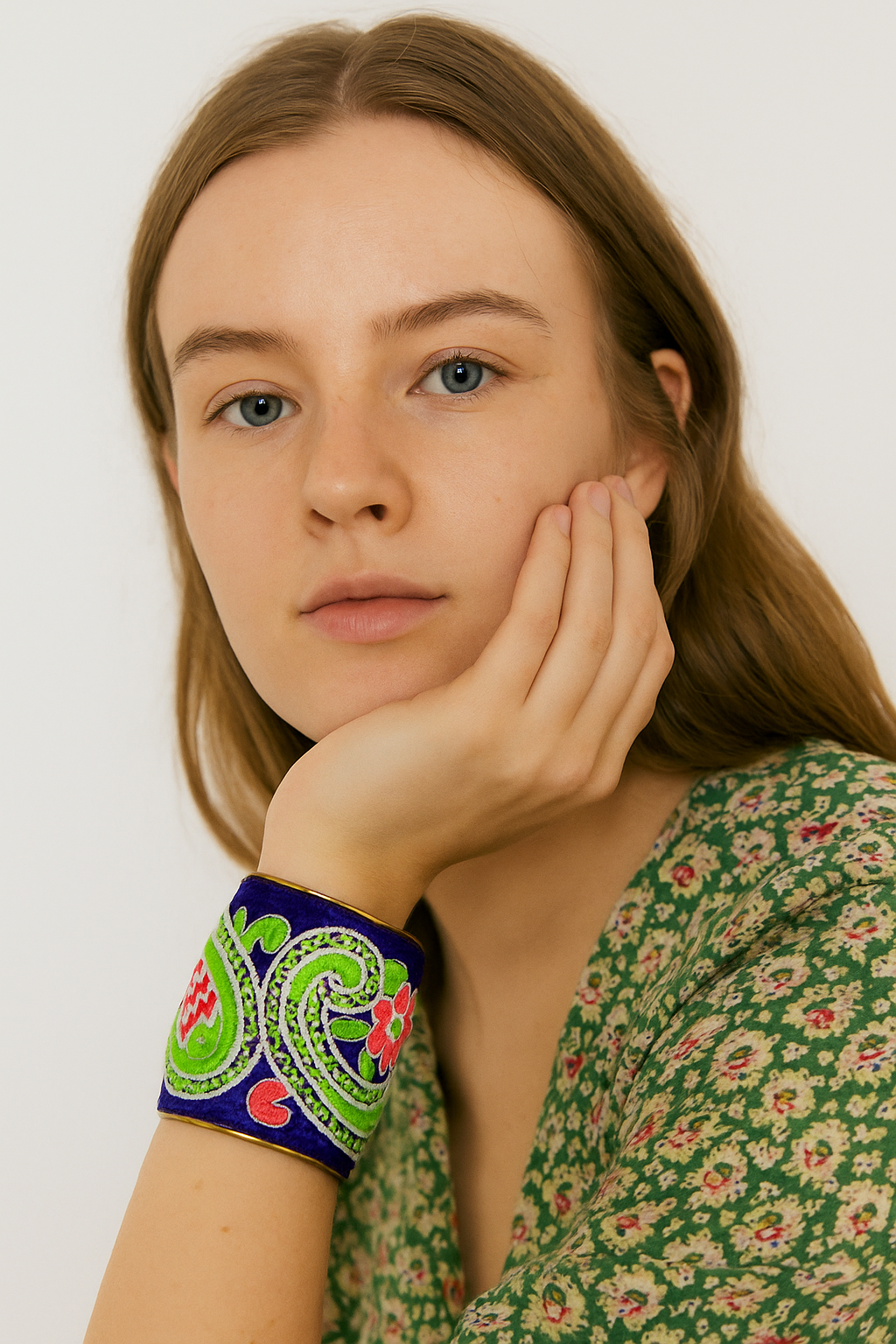 A female model in a green floral blouse rests her chin on her hand, wearing a three-inch brass cuff bracelet wrapped in royal blue velvet with embroidered neon green and coral paisley motifs; the open-back adjustable fit is visible against a white background.