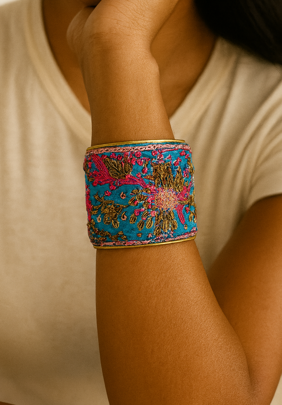 Woman’s wrist wearing the Redd Dott “Shai” 2″ cuff—teal/peacock-blue fabric with multicolor floral embroidery (magenta, gold, green) and subtle metallic threads, framed in brushed brass with an open back.