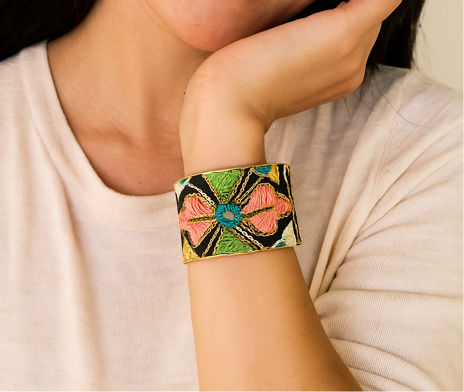 Female model with long dark hair wearing a cream top, resting her cheek on her hand and showcasing a 1-inch wide brass cuff bracelet wrapped in black fabric with multicolor embroidered petals and a turquoise center on her left wrist—a standout example of cuff bracelets for women.