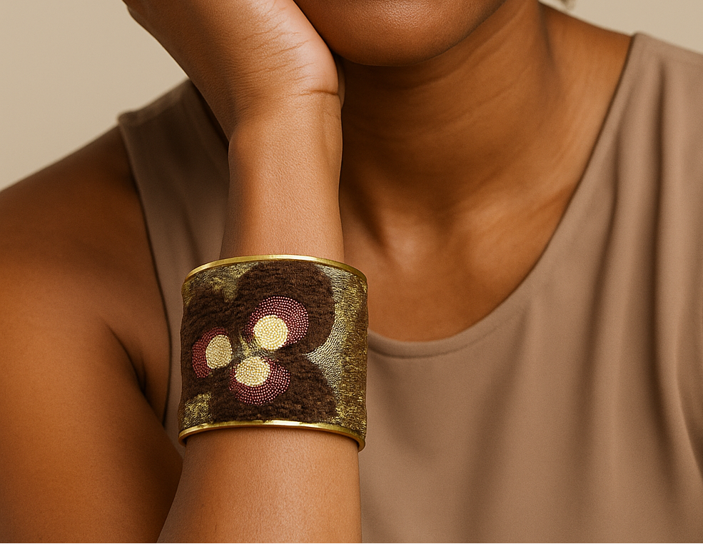 Female model with natural curls in a neutral top, resting her chin on her hand and showcasing a brass cuff bracelet wrapped in brown and gold textured velvet with burgundy and yellow floral embroidery on her left wrist. A standout example of cuff bracelets for women.