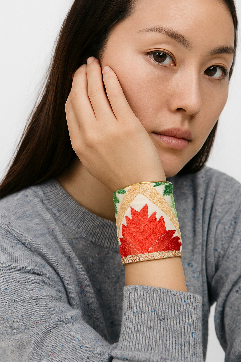 Female model with dark hair wearing a light gray sweater, resting her chin on her hand and showcasing a 3-inch wide brass cuff bracelet wrapped in embroidered ivory fabric with green, yellow, and orange geometric and botanical motifs on her left wrist.