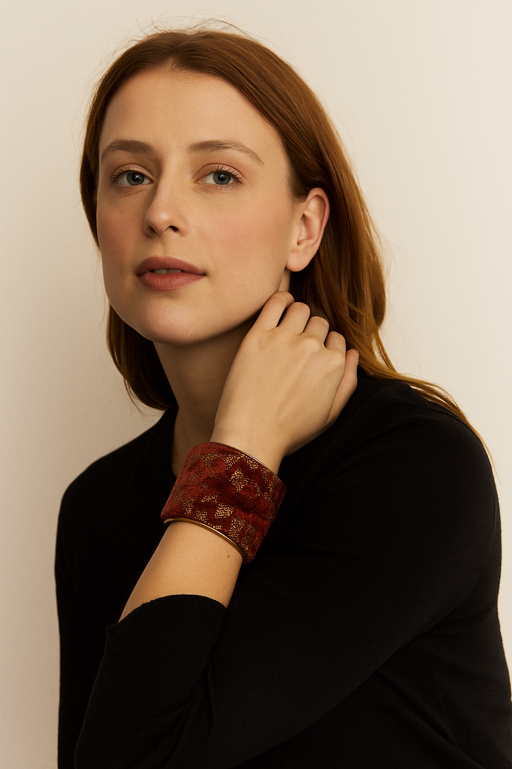 Female model with red hair wearing a black top, resting her chin on her hand and showcasing a 2-inch wide brass cuff bracelet wrapped in rust velvet with a gold geometric medallion on her left wrist. A bold example of cuff bracelets for women.