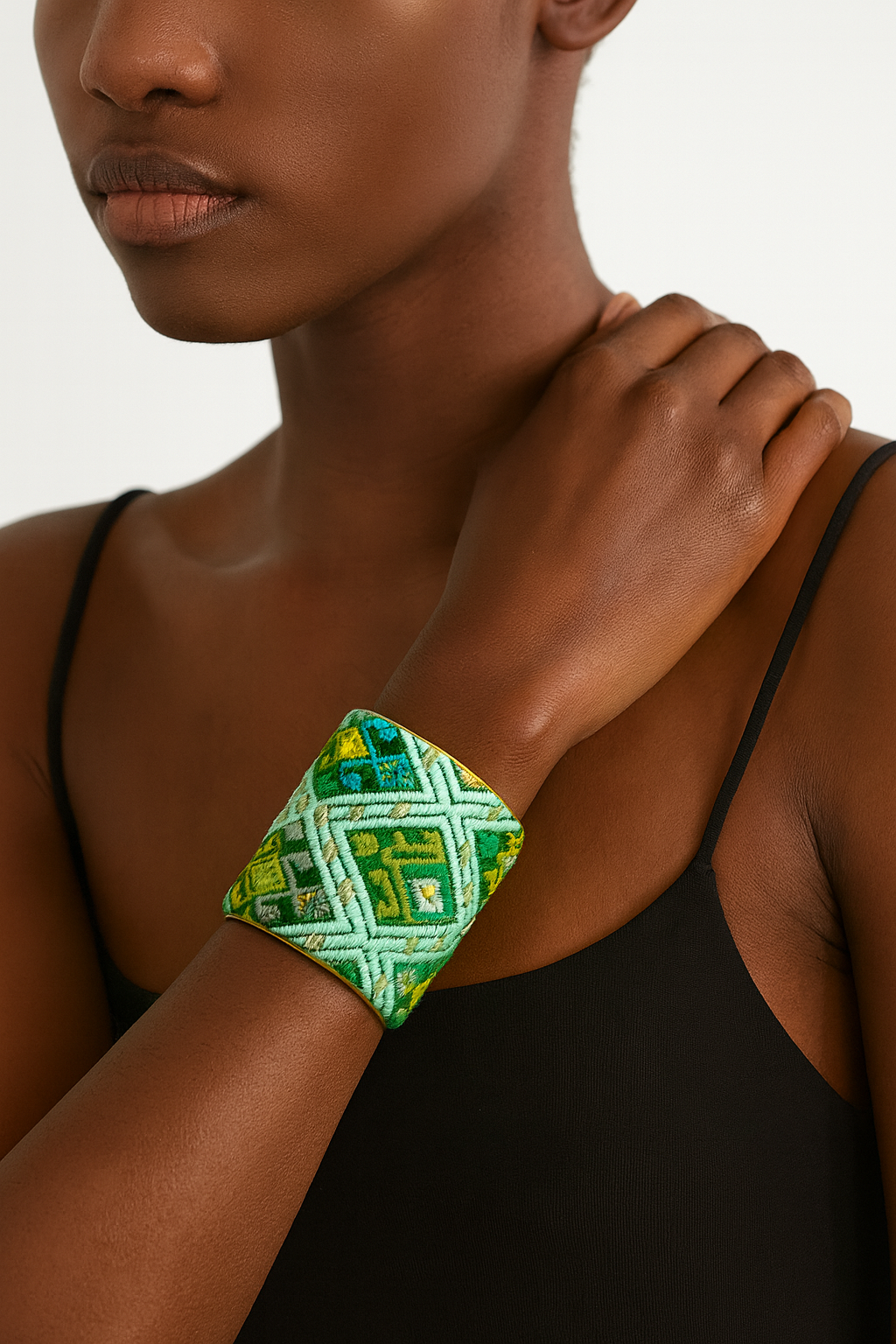 Close-up of a woman’s wrist and lower face, wearing a Redd Dott 3″ wide fabric cuff. The bracelet’s surface features densely stitched geometric diamonds and chevrons in shades of emerald, lime, and mint green, layered over soft cotton and edged with a slim open-back brass frame as her hand gently rests on her shoulder.