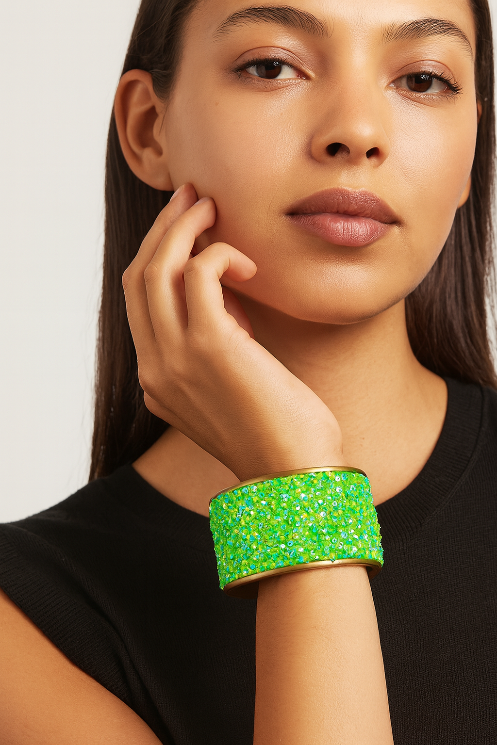 Close-up of a woman’s wrist and lower face, wearing a Redd Dott 3″ wide fabric cuff. The cuff’s surface is densely covered in lime-green sequins and iridescent crystal beads that catch the light, all mounted on a soft cotton base and framed by a slender open-back brass shell as her hand gently touches her cheek.