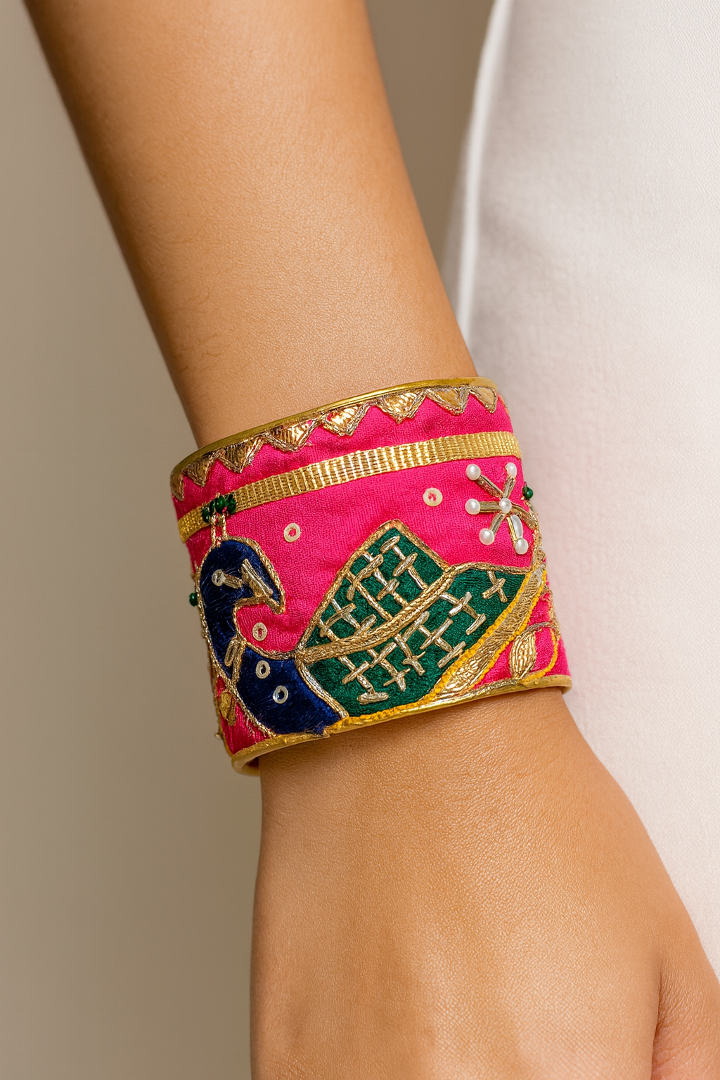 Close-up of the Redd Dott Pea 2-inch cuff bracelet, featuring vibrant pink fabric adorned with a detailed embroidered peacock in blue and green, accented with gold trim and pearl embellishments for an elegant, artistic look.