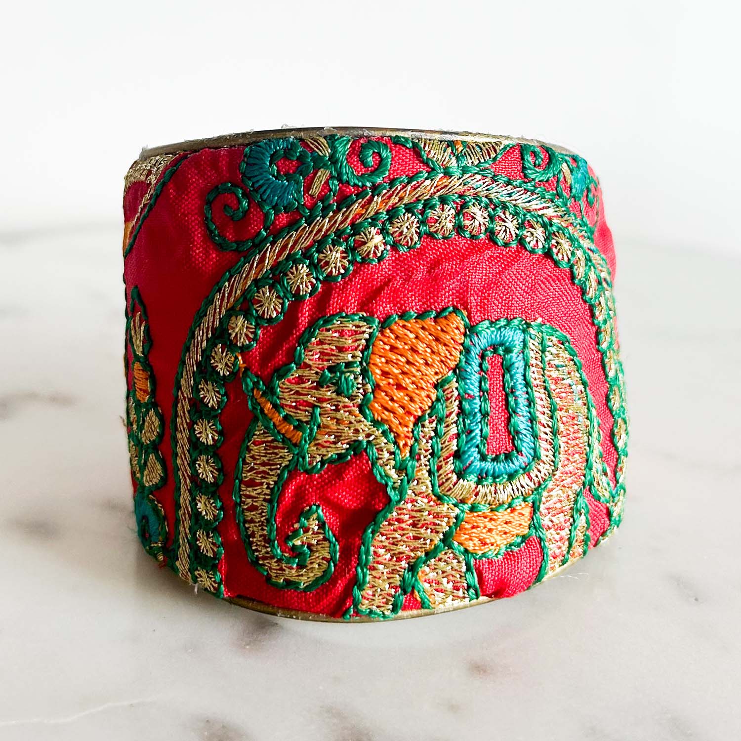 Studio image of the Redd Dott “Zelda” 3″ fabric cuff bracelet resting on a white marble surface. The vibrant red textile showcases a gleaming gold‐threaded elephant motif surrounded by verdant green filament vines and tiny ruby-red accents, with the polished brass interior peeking through the open back.