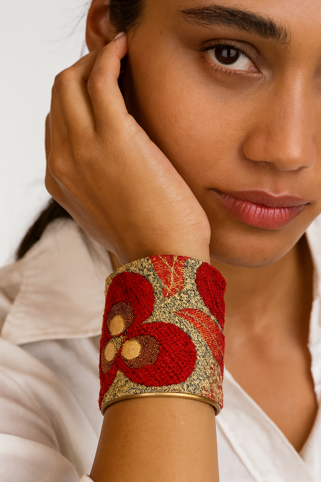 Close-up of a woman’s wrist and lower face, wearing the Redd Dott “Rowan” 3″ wide fabric cuff. Oversized crimson floral blooms with champagne-gold centers and leaves sit on a speckled antique-gold jacquard ground, framed by smooth matte-brass edges with an open-back shell as her hand rests near her cheek.