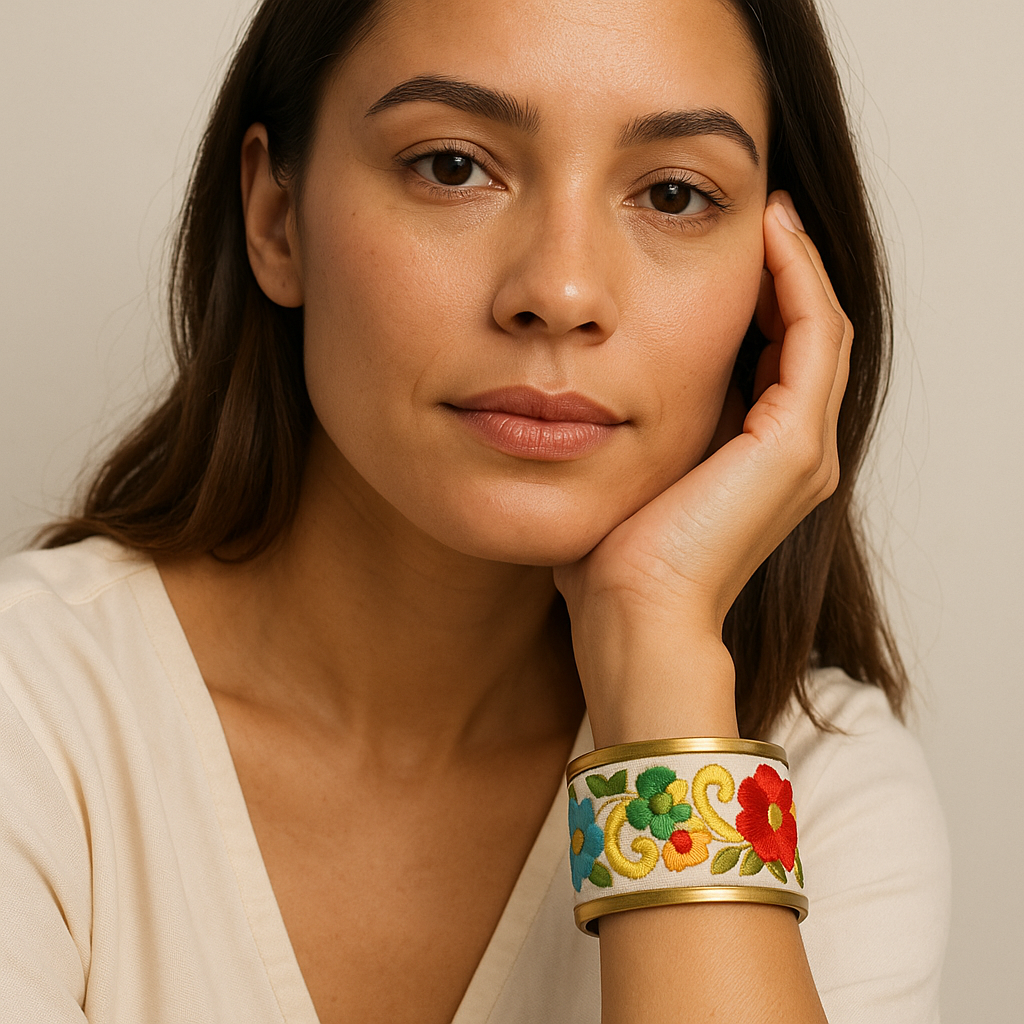 Close-up of a woman resting her face on her hand wearing the Redd Dott “Sadie” 1″ cuff—slim brass-rim bracelet on a soft champagne/ivory ground with bright embroidered florals (red, blue, green) and yellow vine scrolls.