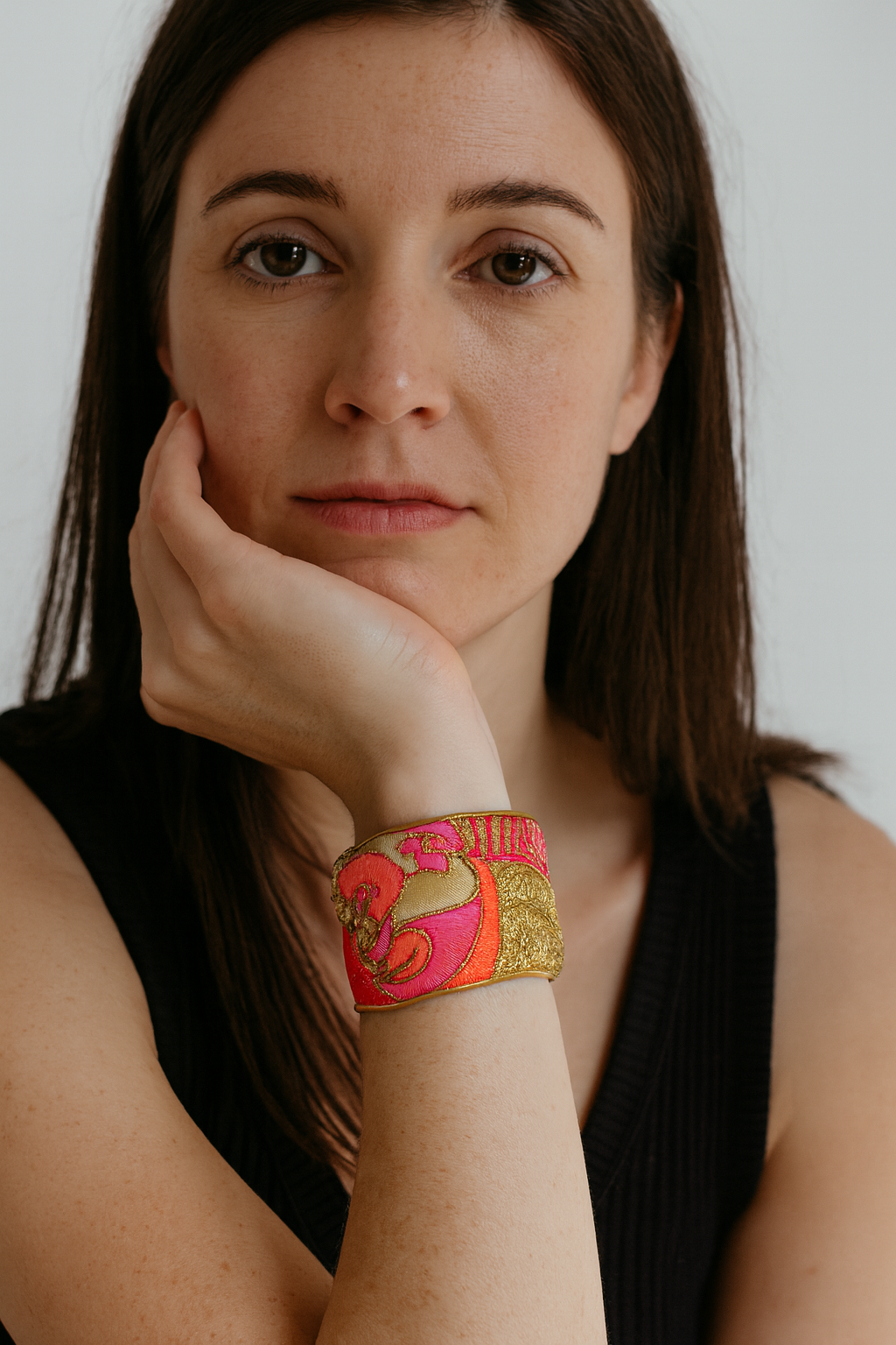 Close-up of a woman resting her chin on her hand wearing the Redd Dott “Sage” 2″ cuff—brass-trimmed bracelet with vivid fuchsia and metallic gold embroidery on a champagne ground, featuring swirling floral/paisley motifs.