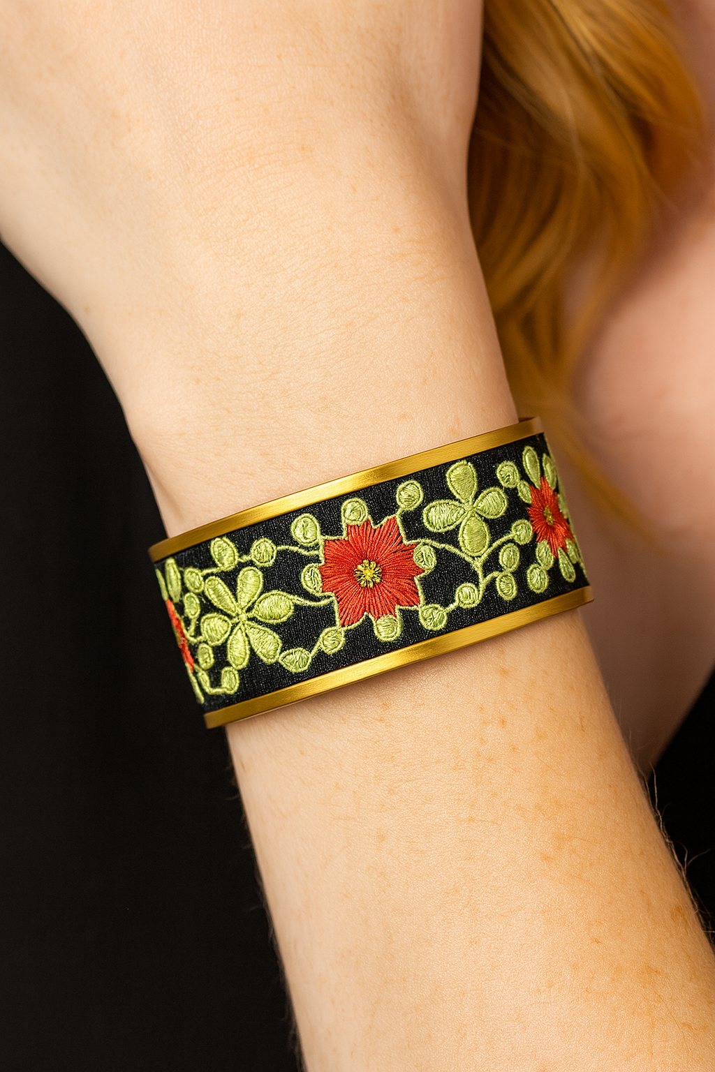 Close-up of a wrist in a black top wearing the Redd Dott “Sammy” 1″ cuff—gold-tone edges around a black ground with lime-green embroidered vine florals and bold orange-red starburst blooms.