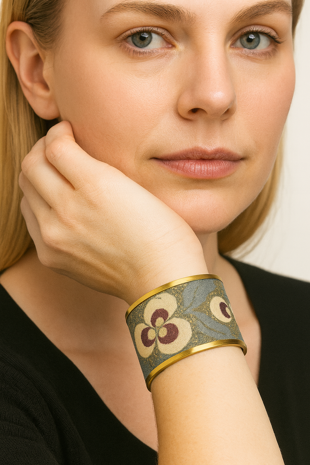 Close-up of a woman in a black top resting her chin on her hand, wearing the Redd Dott “Saylor” 2″ cuff—antique-brass edges framing a slate-blue jacquard band with ivory florals (plum centers) and olive leaf vines.