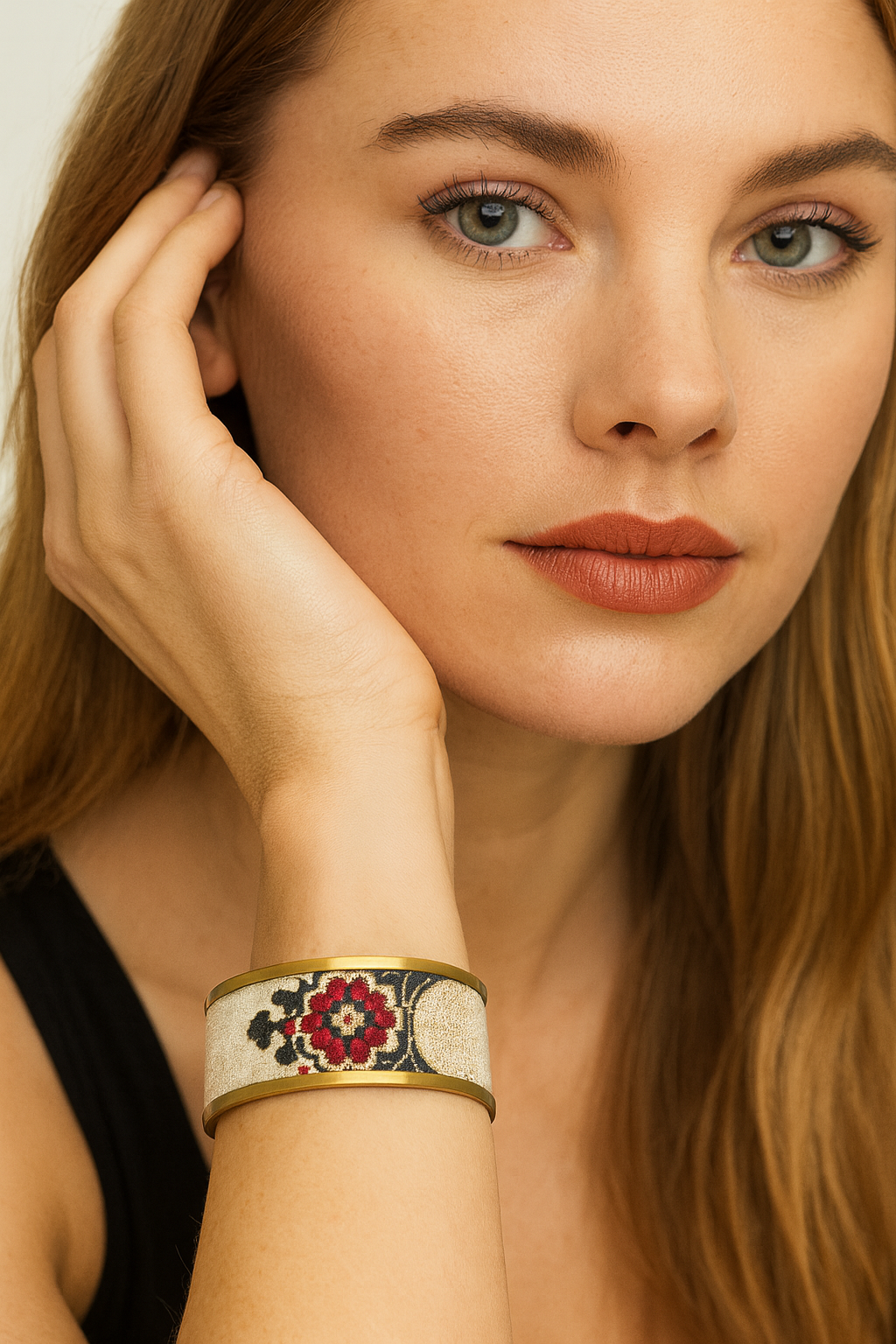Close-up of a woman in a black tank resting her cheek on her hand, wearing the Redd Dott “Scarlet” 1″ cuff—matte-brass edges framing an ivory brocade band with a red rosette medallion and black scrollwork.