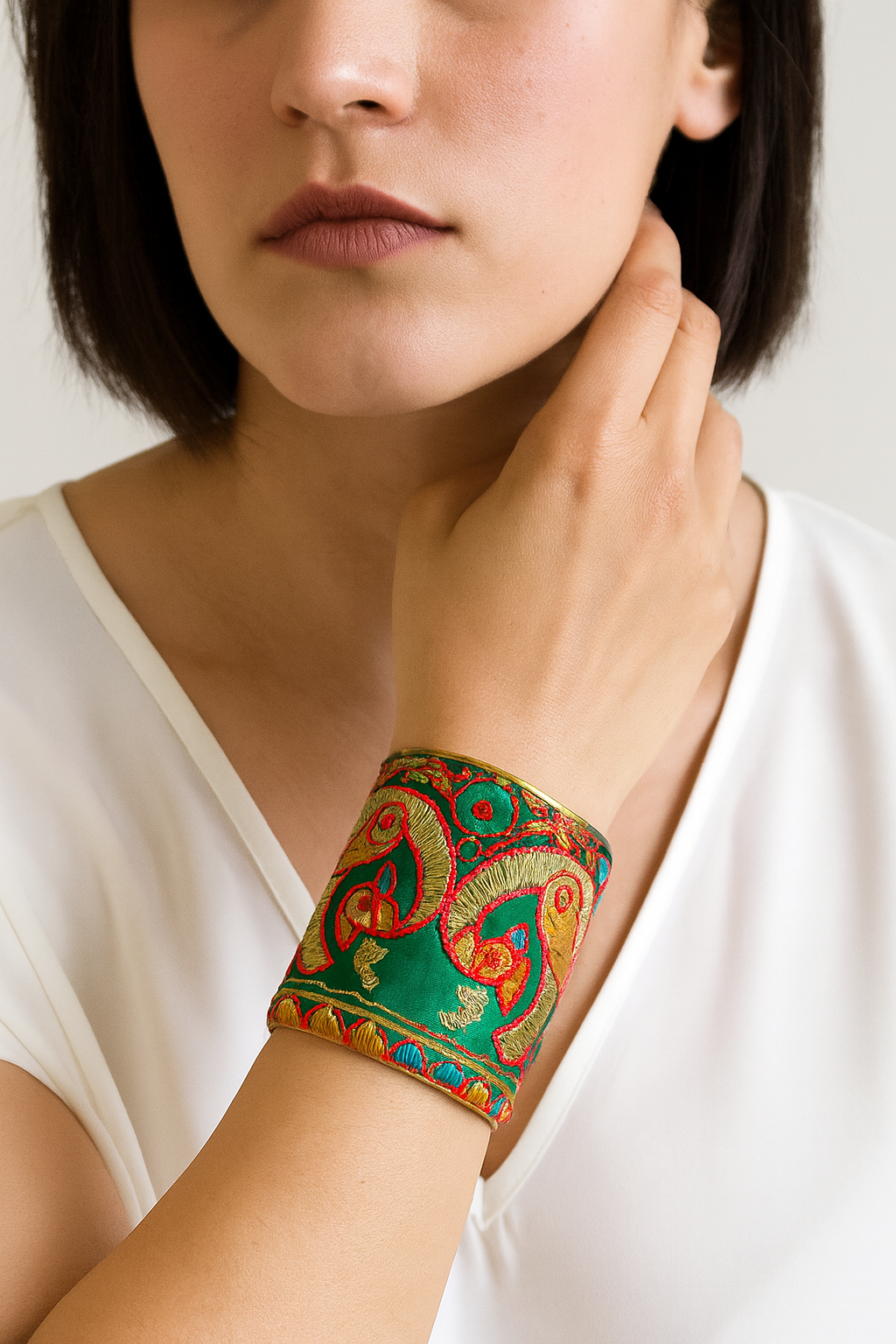 Close-up of a woman wearing the Redd Dott “Seraphina” 3″ cuff—emerald-green satin with ornate gold embroidery and red-outlined paisley/scroll motifs, finished with antique-brass edges and a multicolor scalloped border.