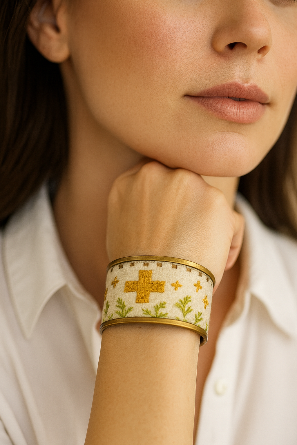 Woman’s wrist wearing the Redd Dott “Shoda” 1″ cuff—ivory fabric with mustard-gold cross motifs, tiny plus accents, and a border of olive-green leafy sprigs, framed by brushed-brass rims with an open back.