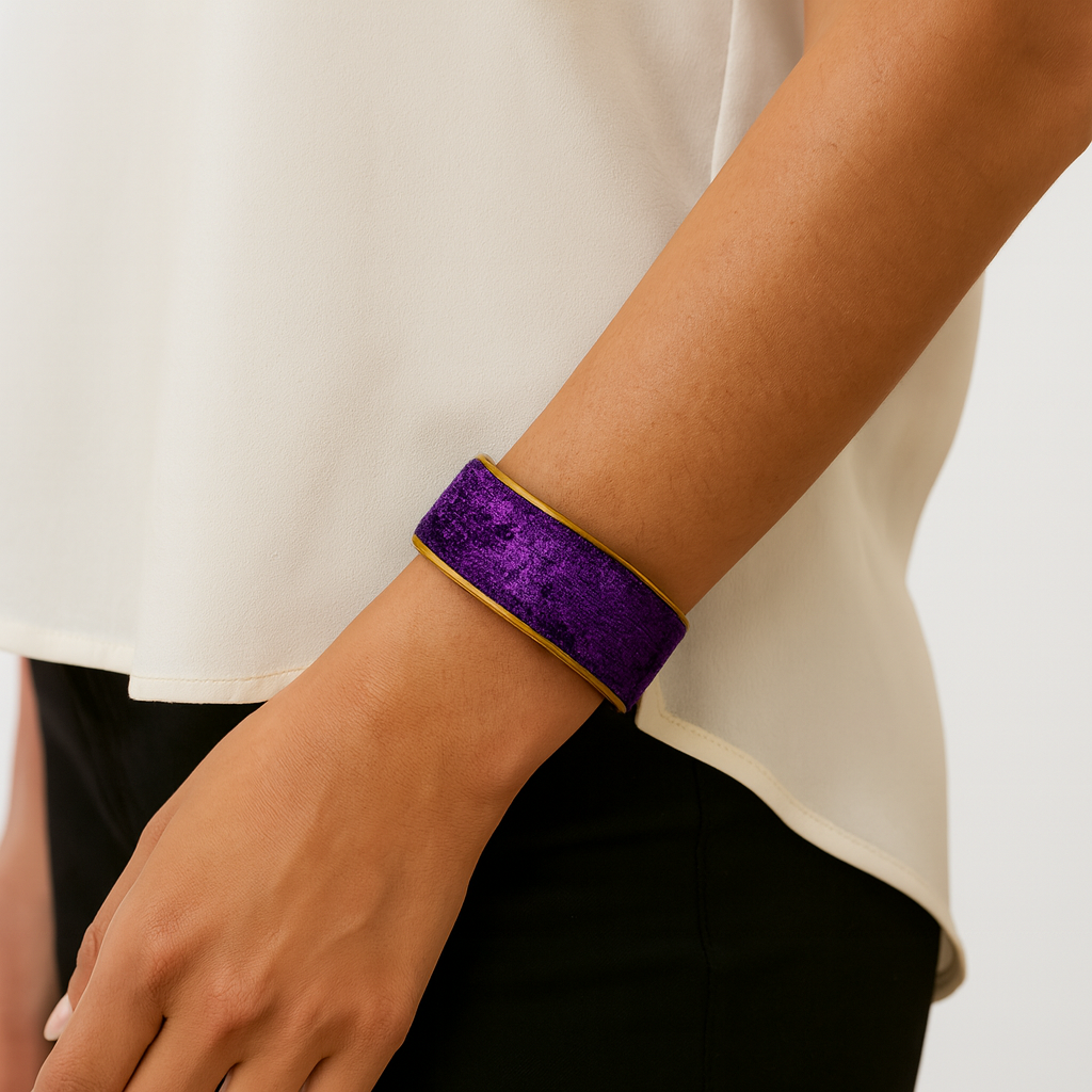 Woman’s wrist wearing the Redd Dott “Sigal” 1″ cuff—sleek, royal-purple crushed velvet with a subtle sheen, edged in slim brushed-brass rims and an open back; styled with a cream blouse and black pants.