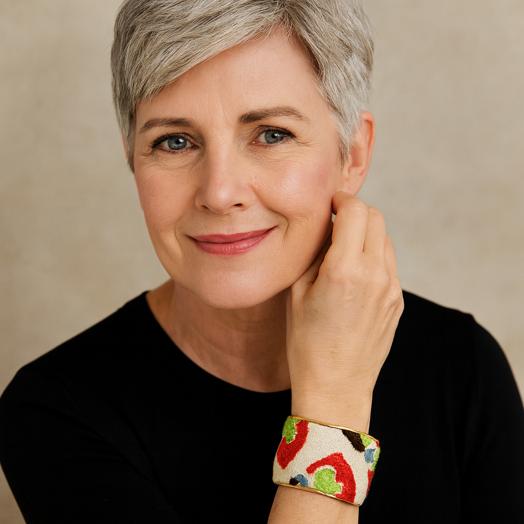 Close-up of a woman’s wrist and lower face, wearing the Redd Dott “Silvya” 2″ wide fabric cuff. The bracelet features a sandy-ivory canvas scattered with painterly poppy-red crescents, leaf-green dabs, and midnight-blue accents, all framed by slim polished-brass edges and an open-back shell as her hand rests near her cheek.