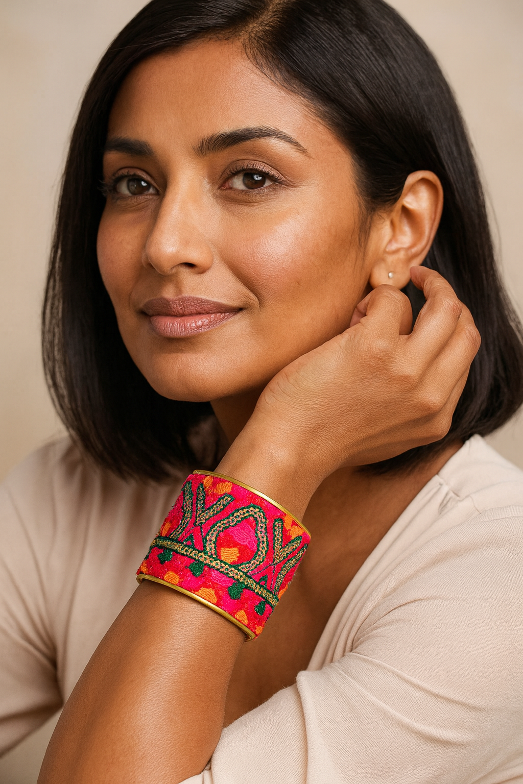 Close-up of a woman’s wrist and lower face, wearing the Redd Dott “Stefania” 2″ wide fabric cuff. The electric fuchsia band is densely embroidered in emerald and chartreuse threads forming bold X-O motifs and chevron shapes, finished with a tiny scalloped dot border and framed by a slim open-back brass shell as her hand rests near her ear.