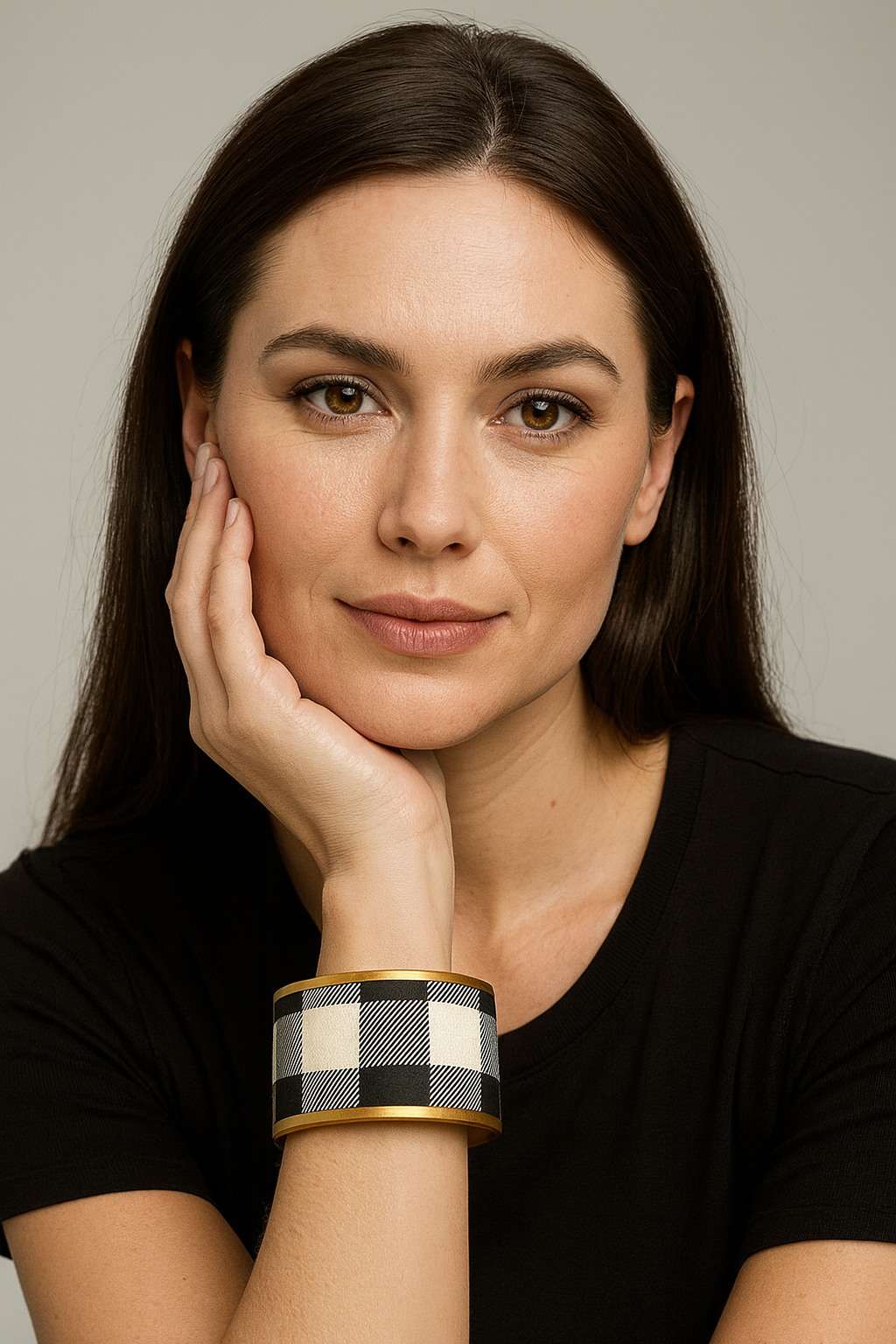 Close-up of a woman’s wrist and lower face, wearing the Redd Dott “Sutton” 2″ wide fabric cuff. The bracelet showcases classic black-and-ivory buffalo check plaid with visible twill texture, framed by smooth matte-brass edges and an open-back shell as her hand rests at her cheek.