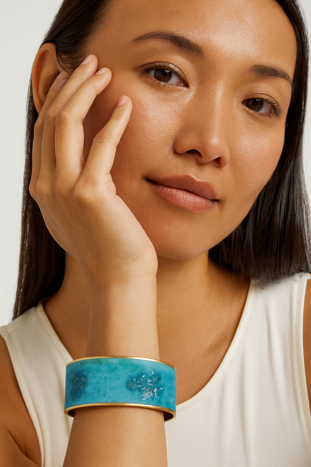 Close-up of a woman’s wrist and lower face, wearing the Redd Dott “Sylas” 1″ wide fabric cuff. The slim bracelet features a lagoon-blue/teal watercolor wash with soft marbling and scattered silver flecks, framed by slender polished-brass edges and an open-back shell as her hand rests near her cheek.