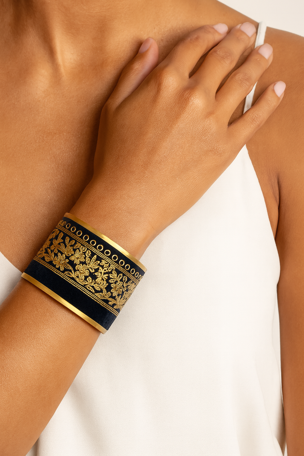 Close-up of a woman’s wrist and forearm, wearing the Redd Dott “Terra” 2″ wide fabric cuff. The deep midnight-navy velvet band is detailed with ornate metallic-gold floral borders and a row of tiny gold rings, framed by smooth matte-brass edges and an open-back shell as her hand rests near her collarbone.