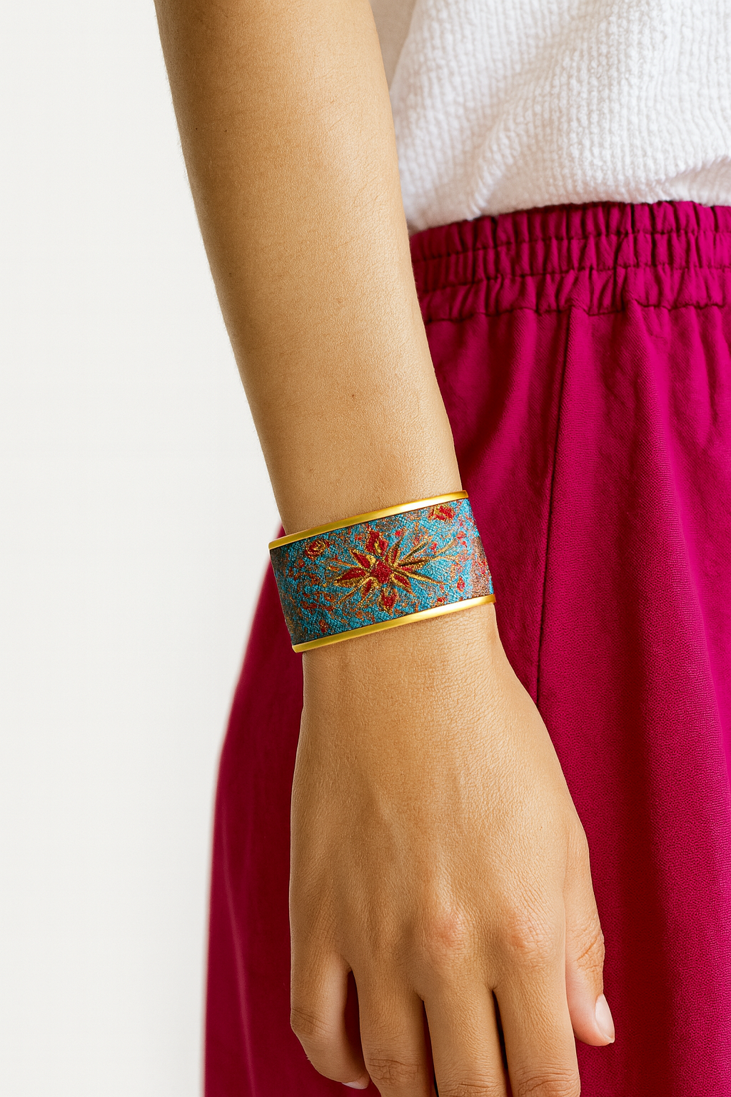 Close-up of a woman’s wrist wearing the Redd Dott “Tina” 1″ wide fabric cuff. The slim teal jacquard band features a red-and-gold starburst medallion with tiny filigree accents, framed by polished brass edges and an open-back shell as her hand rests near fuchsia trousers.