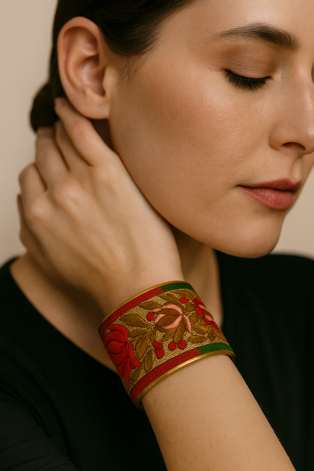 Close-up of a woman’s wrist and lower face, wearing the Redd Dott “Tivona” 3″ wide fabric cuff. The bracelet features holiday-rich embroidery—rosy pink and deep red blossoms, scarlet berries, and antique-gold leaves—on a warm red ground with slim green and red borders, all framed by a smooth open-back brass shell as her hand rests near her ear.