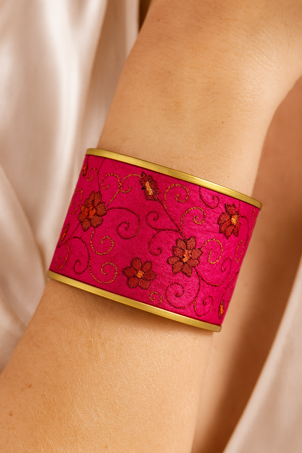 Close-up of a woman’s wrist wearing the Redd Dott “Tora” 2″ wide fabric cuff. The bold fuchsia satin band is embroidered with shimmering gold scroll vines and copper-orange floral blossoms, framed by smooth matte-brass edges with an open-back shell.