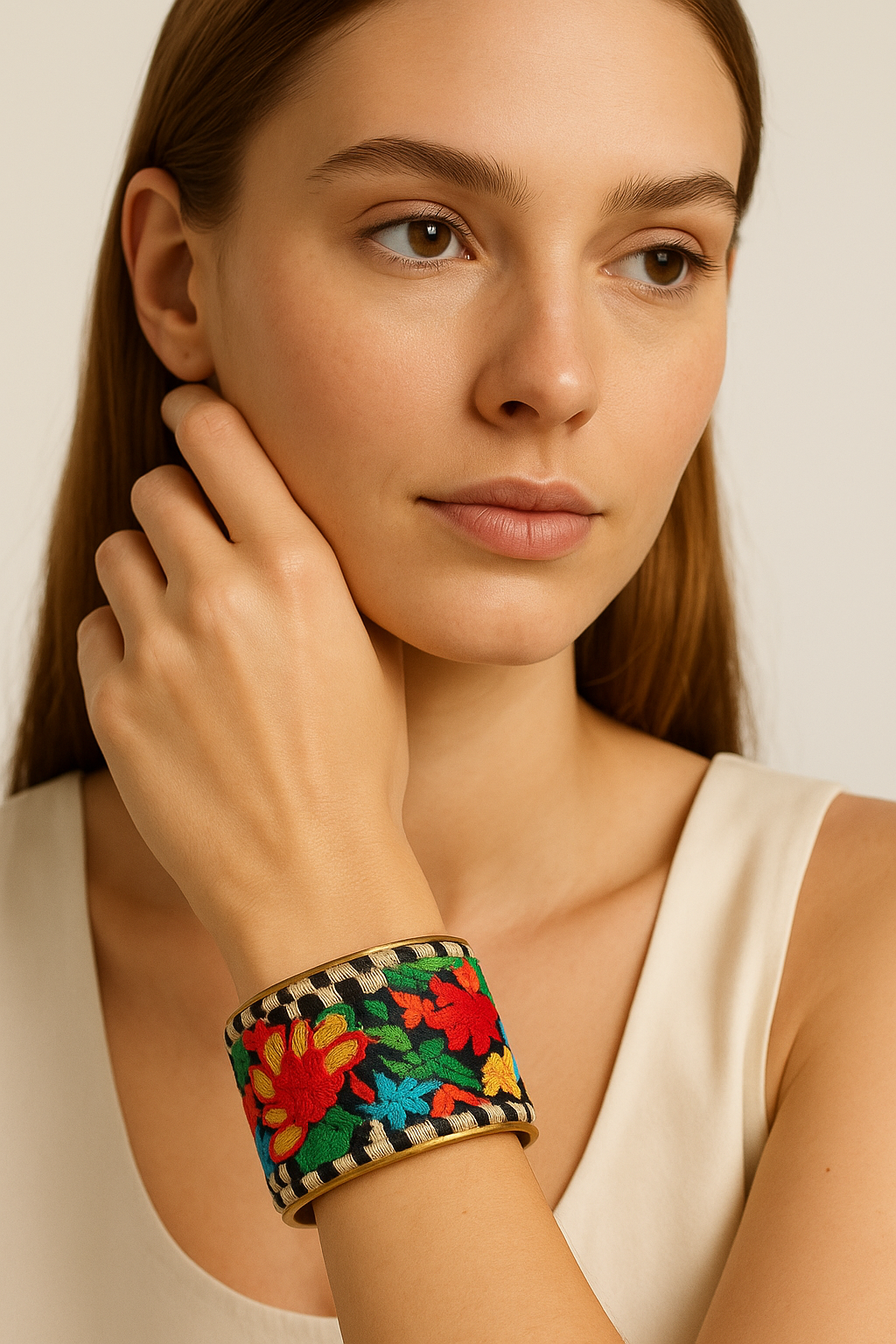 Close-up of a woman’s wrist and lower face, wearing the Redd Dott “Tropica” 2″ wide fabric cuff. The bracelet shows saturated tropical florals—scarlet, marigold, turquoise, and leaf green—embroidered on a black ground, bordered by black-and-ivory check trim and framed in a slim open-back brass shell as her hand rests near her cheek.