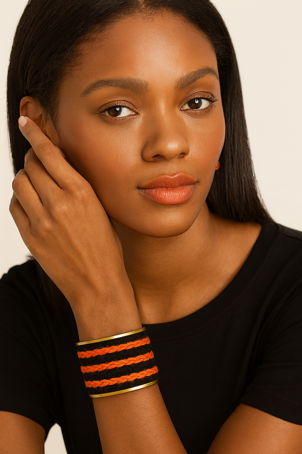 Close-up of a woman’s wrist and lower face, wearing the Redd Dott “Tyler” 2″ wide fabric cuff. The matte-black wool band is wrapped with three chunky, hand-braided orange stripes running horizontally, bordered by smooth polished-brass edges and an open-back shell as her hand brushes her hair.