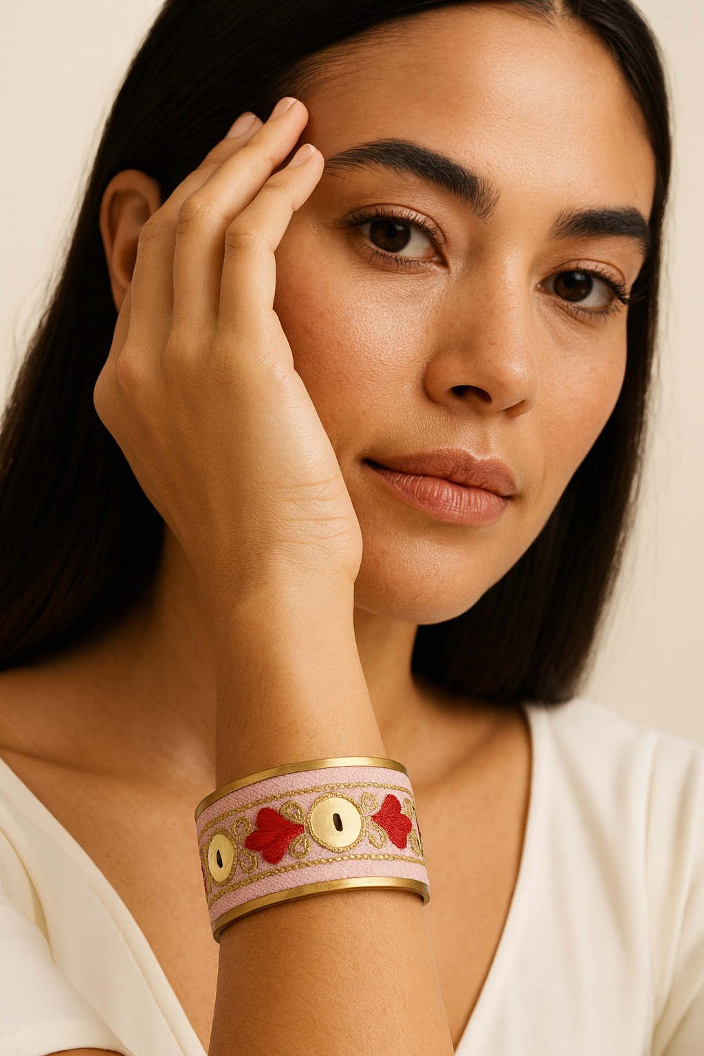 Close-up of a woman’s wrist and lower face, wearing the Redd Dott “Valentina” 1″ wide fabric cuff. The blush-pink textile band is trimmed with matte-brass edges and embellished with crimson heart/clover embroidery and round gold medallion discs linked by champagne-gold filigree stitching, shown with an open-back frame as her hand rests near her temple.