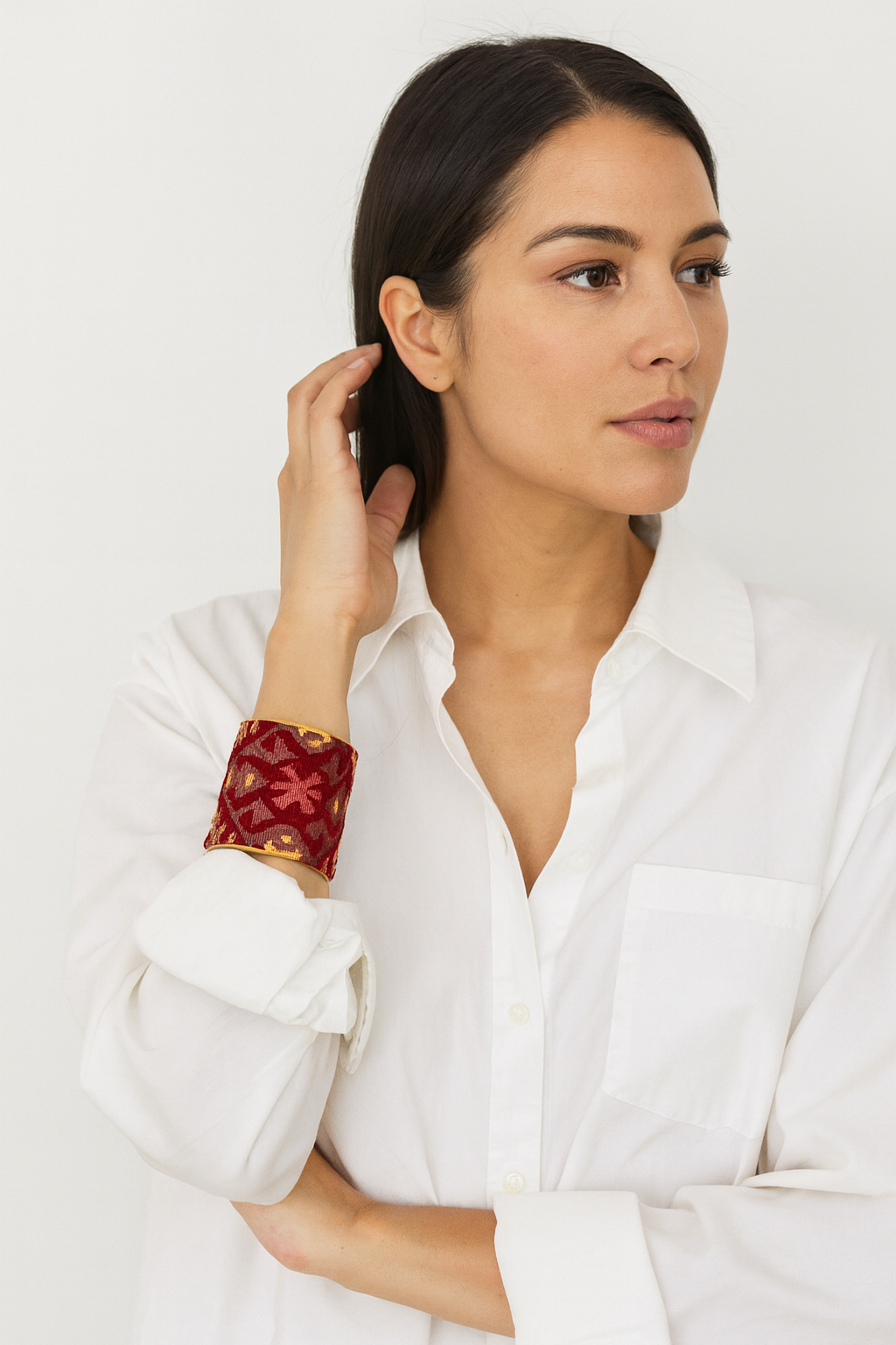 Close-up of a woman’s wrist and lower face, wearing the Redd Dott “Valentine” 3″ wide fabric cuff. The rich crimson cotton base is overlaid with intricate metallic-gold brocade-like floral and geometric motifs in a velvety relief, set within a gently curved open-back brass shell as her hand brushes back her hair.