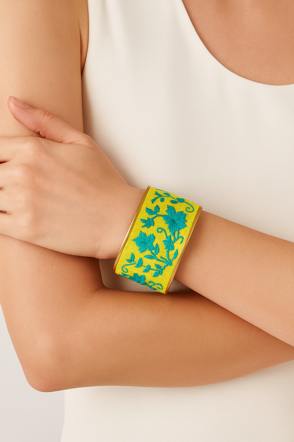 Close-up of a woman’s wrist and forearm, wearing the Redd Dott “Veda” 2″ wide fabric cuff. The bright chartreuse-green cotton cuff is embroidered with swirling teal floral vines and blossoms, all nestled within a slim open-back polished brass frame as her other hand lightly grips her opposite elbow.