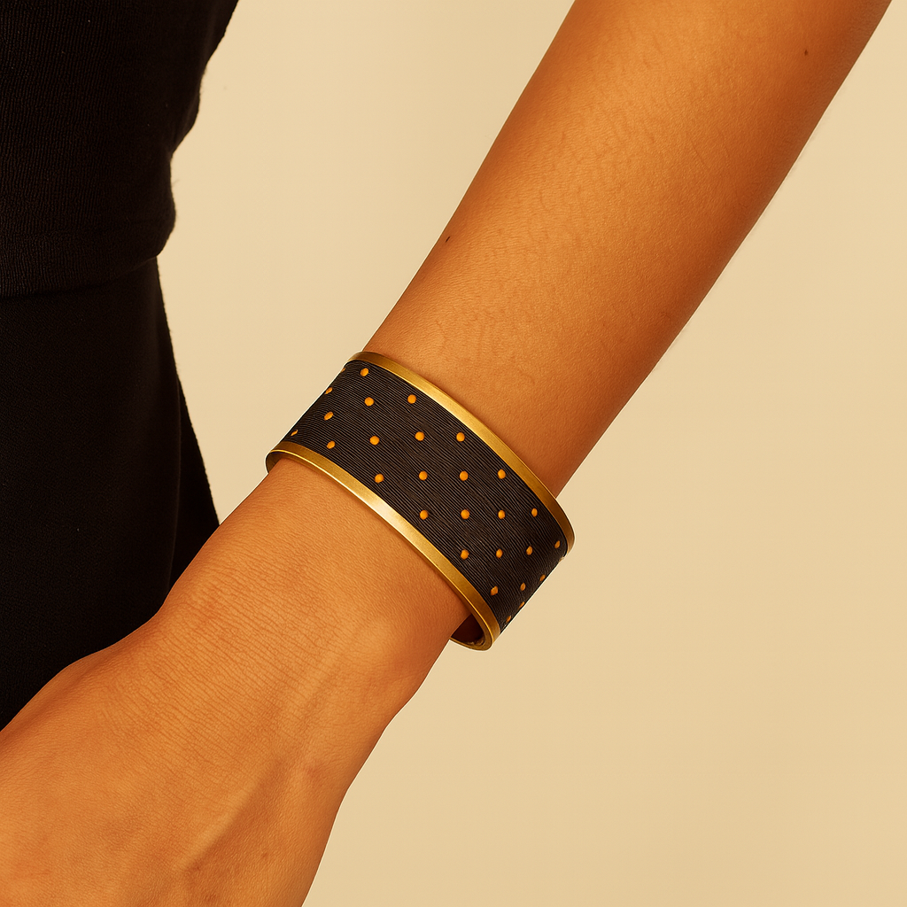 Close-up of a woman’s wrist, wearing the Redd Dott “Vega” 1″ wide fabric cuff. The narrow black grosgrain ribbon is dotted with tiny raised orange stitch-dots in a scattered polka-dot pattern, all framed by a slender open-back brass shell.