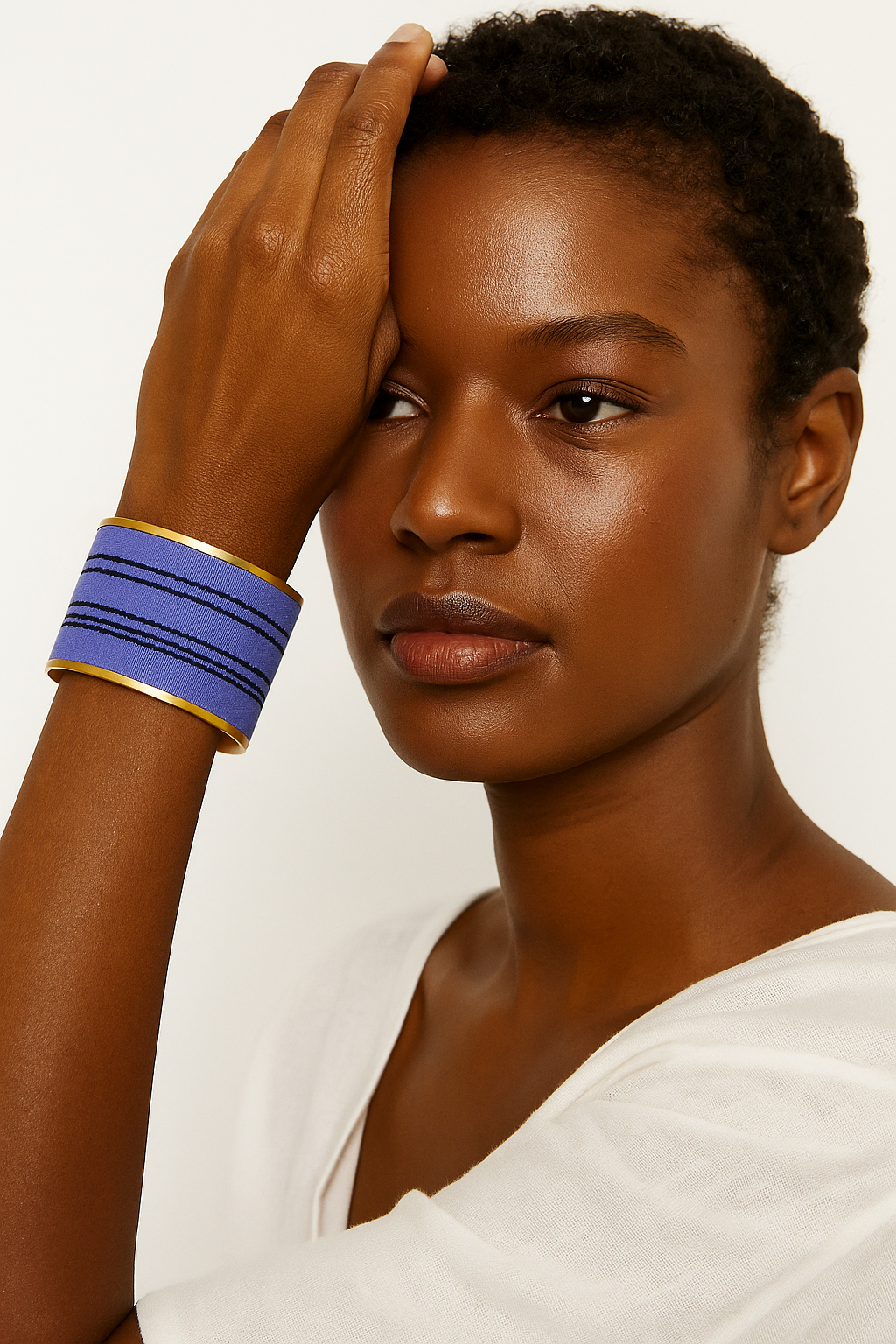 Close-up of a woman’s wrist and lower face, wearing the Redd Dott “Violet” 2″ wide fabric cuff. The vibrant violet cotton band is detailed with three uneven, hand-stitched black thread lines running horizontally, all framed by a slim open-back brass shell as her hand rests against her head.