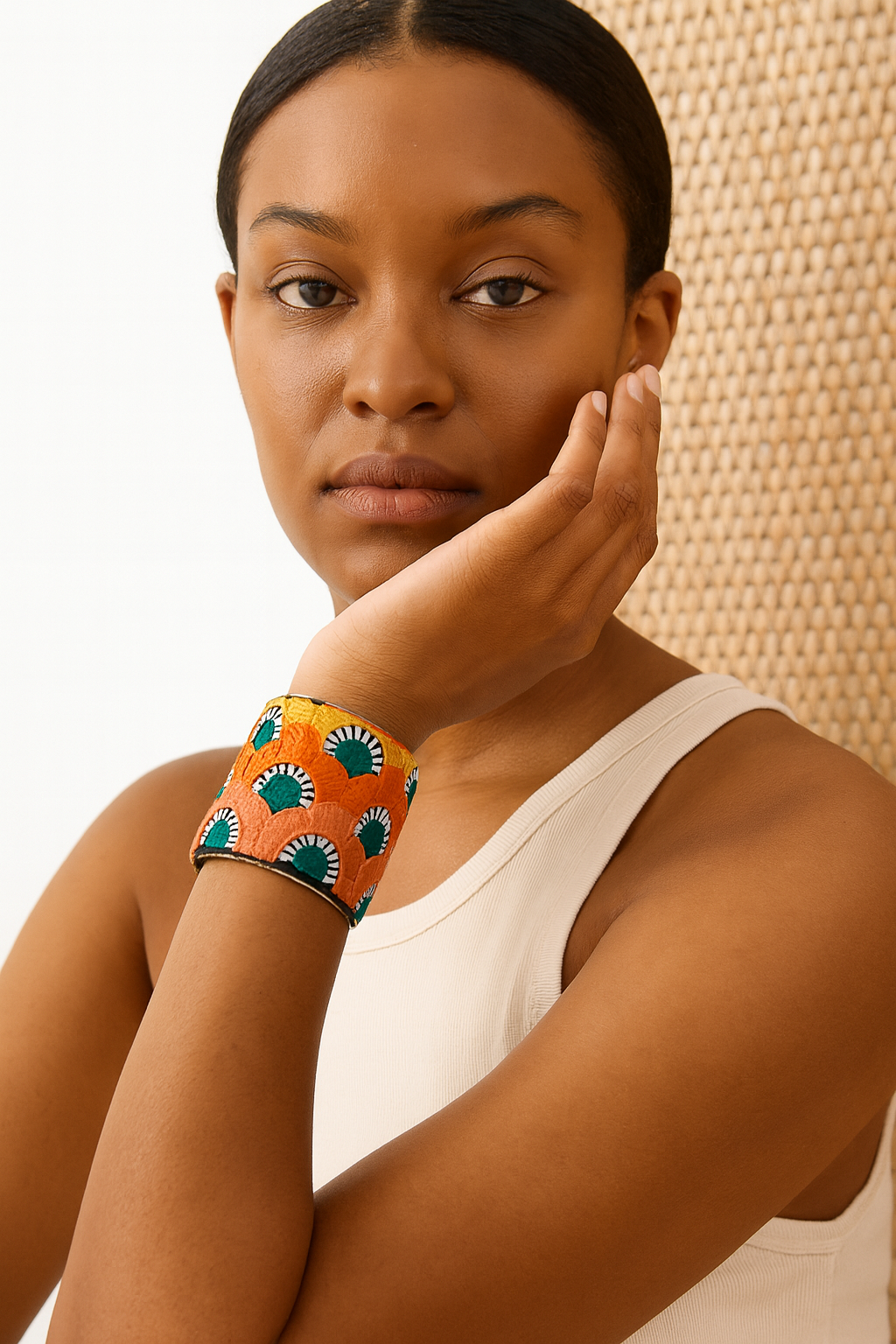 Close-up of a woman’s wrist and lower face, wearing the Redd Dott “Willow” 2″ wide fabric cuff. The vibrant tangerine cotton is embroidered in repeating scallop motifs—each arch in sunshine yellow, ivory, and emerald green with fine black stitch-ray detailing—nestled within a slender open-back brass frame as her hand gently rests against her cheek.