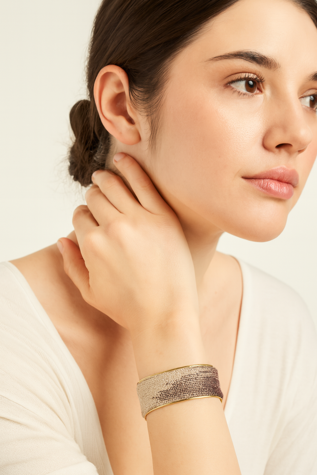 Close-up of a woman’s wrist and lower face, wearing the Redd Dott “Wisteria” 1″ wide fabric cuff. The slim bracelet’s pale lavender cotton is hand-stitched with densely packed silver-metallic thread that fades into smoky charcoal pixels at one edge, all framed by a delicate open-back brass shell as her hand lightly touches her ear.
