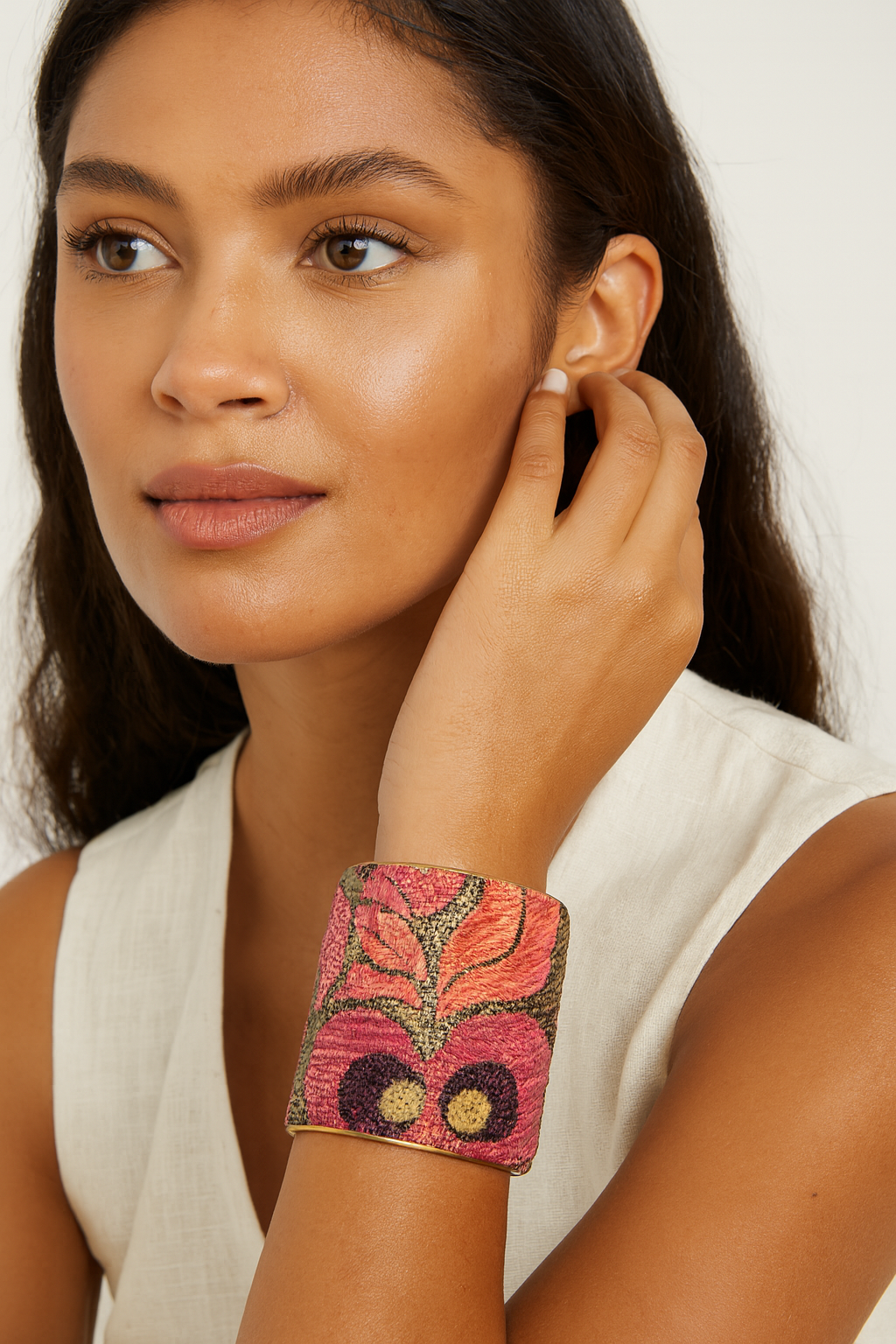 Close-up of a woman’s wrist and lower face, wearing the Redd Dott “Wren” 2″ wide fabric cuff. The bracelet’s textured olive-brown cotton is embroidered with bold coral-pink and magenta floral silhouettes and delicate gold-thread accents, all nestled within a slender open-back brass shell as her hand rests gently near her ear.