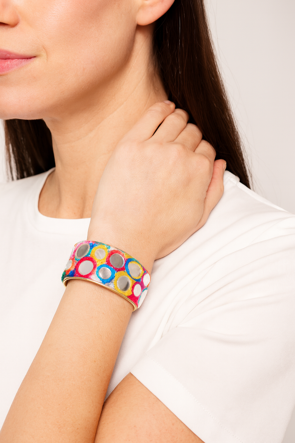 Close-up of a woman’s wrist and lower face, wearing the Redd Dott “Xenia” 1″ wide fabric cuff. The slender white cotton band is hand-painted with glossy multicolor circles—bright pink, cobalt blue, sunshine yellow and metallic silver—each accented with small mirror insets, all framed by a polished brass open-back shell as her hand rests lightly on her shoulder.