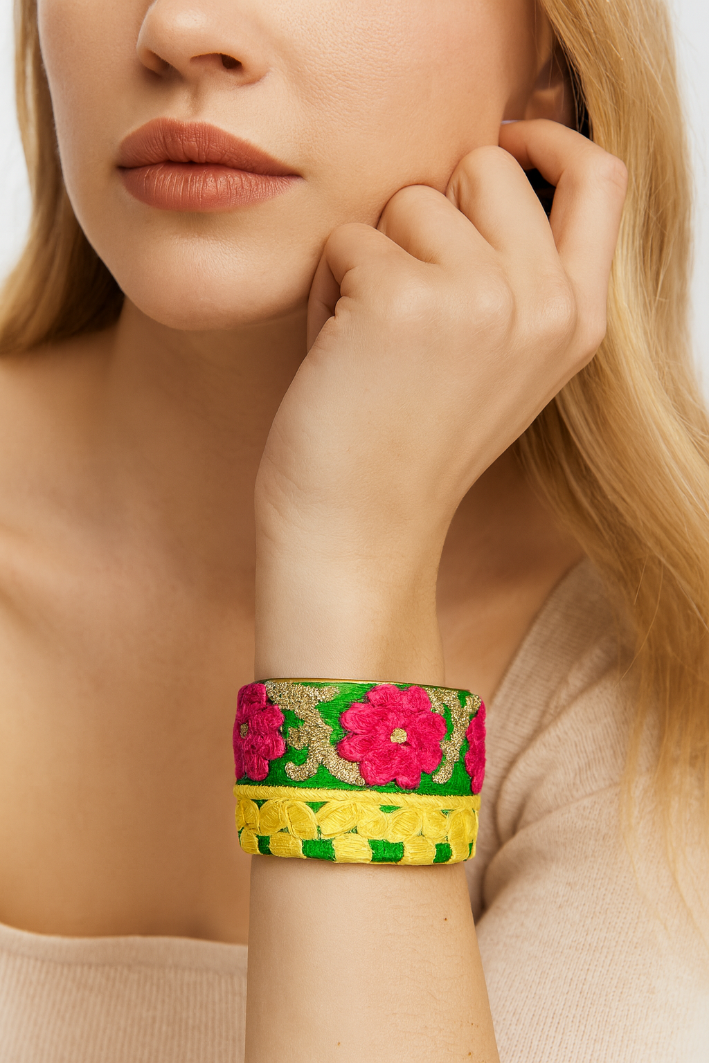 Close-up of a woman’s wrist and lower face, wearing the Redd Dott “Yesonia” 2″ wide fabric cuff. The top half features fuchsia-pink embroidered flowers and glittering metallic-gold scrollwork on vivid emerald-green cotton, while the bottom half shows chunky yellow woven cotton braids, all framed in an open-back brass shell as her hand lightly touches her cheek.