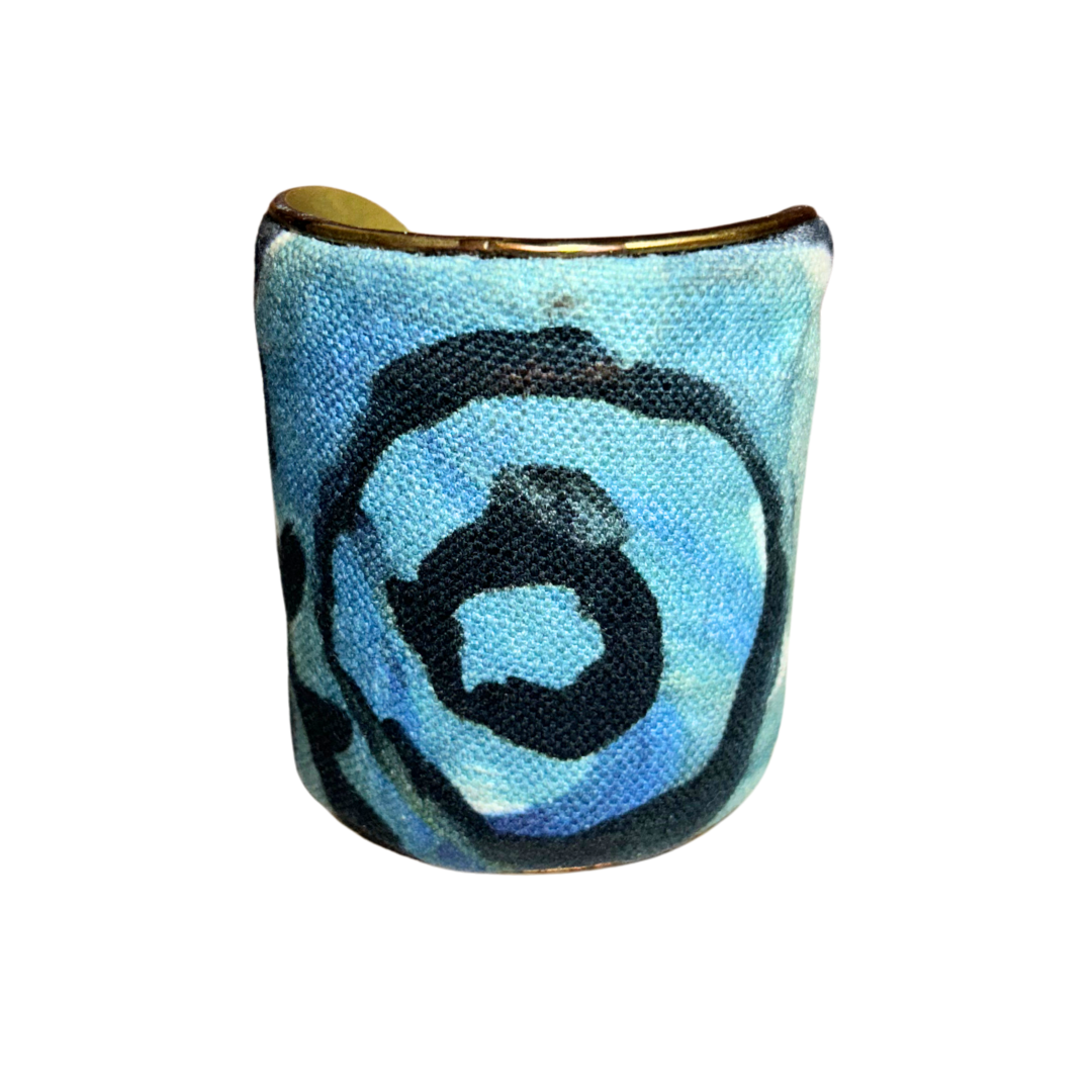 A handmade brass cuff bracelet from Redd Dott Jewelry, crafted and stamped in Shreveport, LA. Features painted blue and white fabric with black abstract circular patterns over a vintage brass base—an expressive piece of inspirational jewelry.